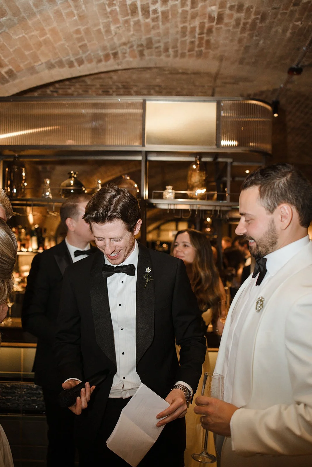 A man wearing a tuxedo with a flower on lapel smiling and looking down at a piece of paper, holding a microphone in one hand, at a formal event with other dressed-up guests in the background.