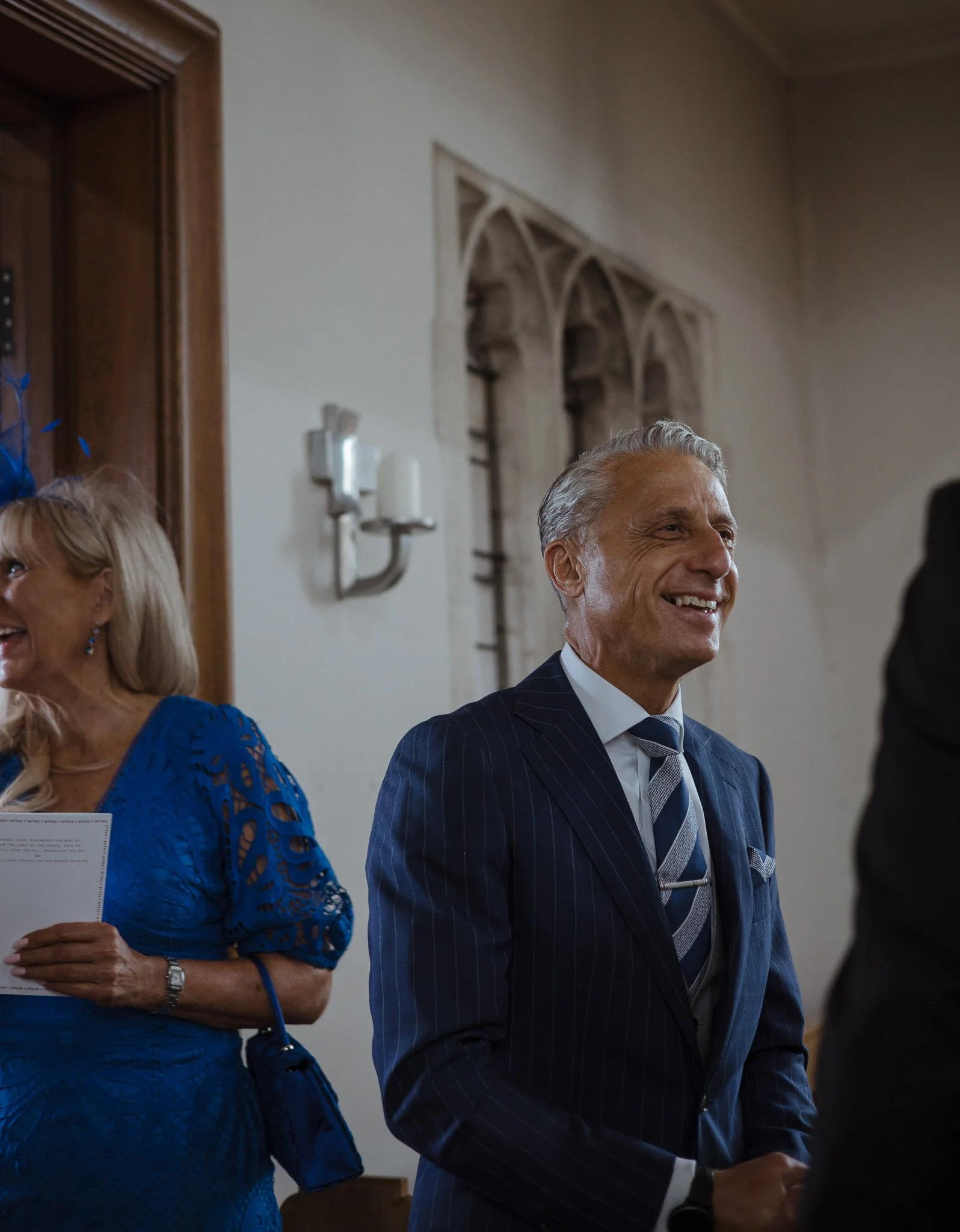 A man smiling in a dark blue pinstripe suit at an indoor gathering with other people, including an older woman in a vibrant blue dress, in front of a wall with stone architectural details.