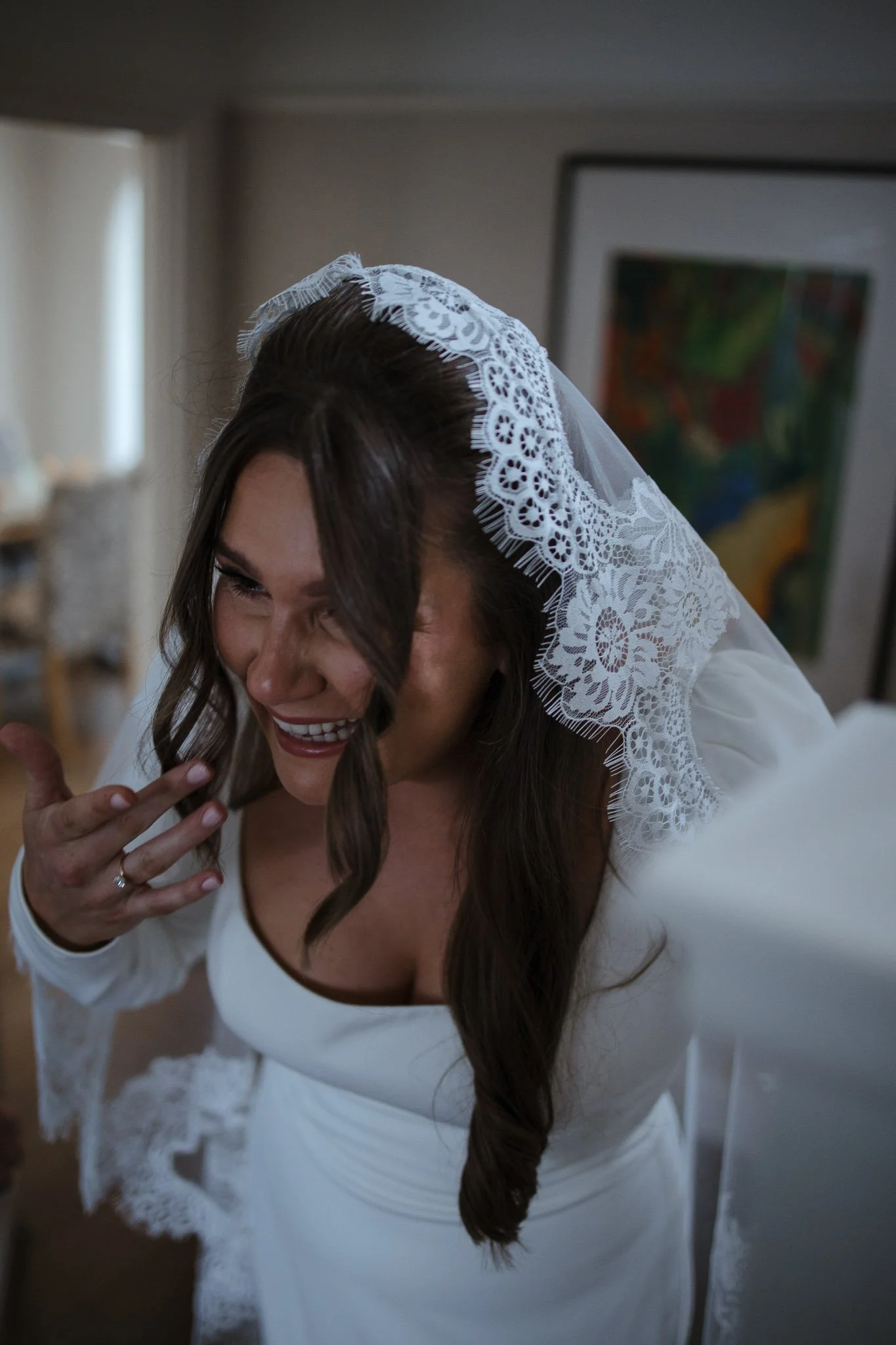A smiling woman in a white dress with lace details, wearing a lace veil, is getting ready for a wedding, standing indoors in front of a framed painting.