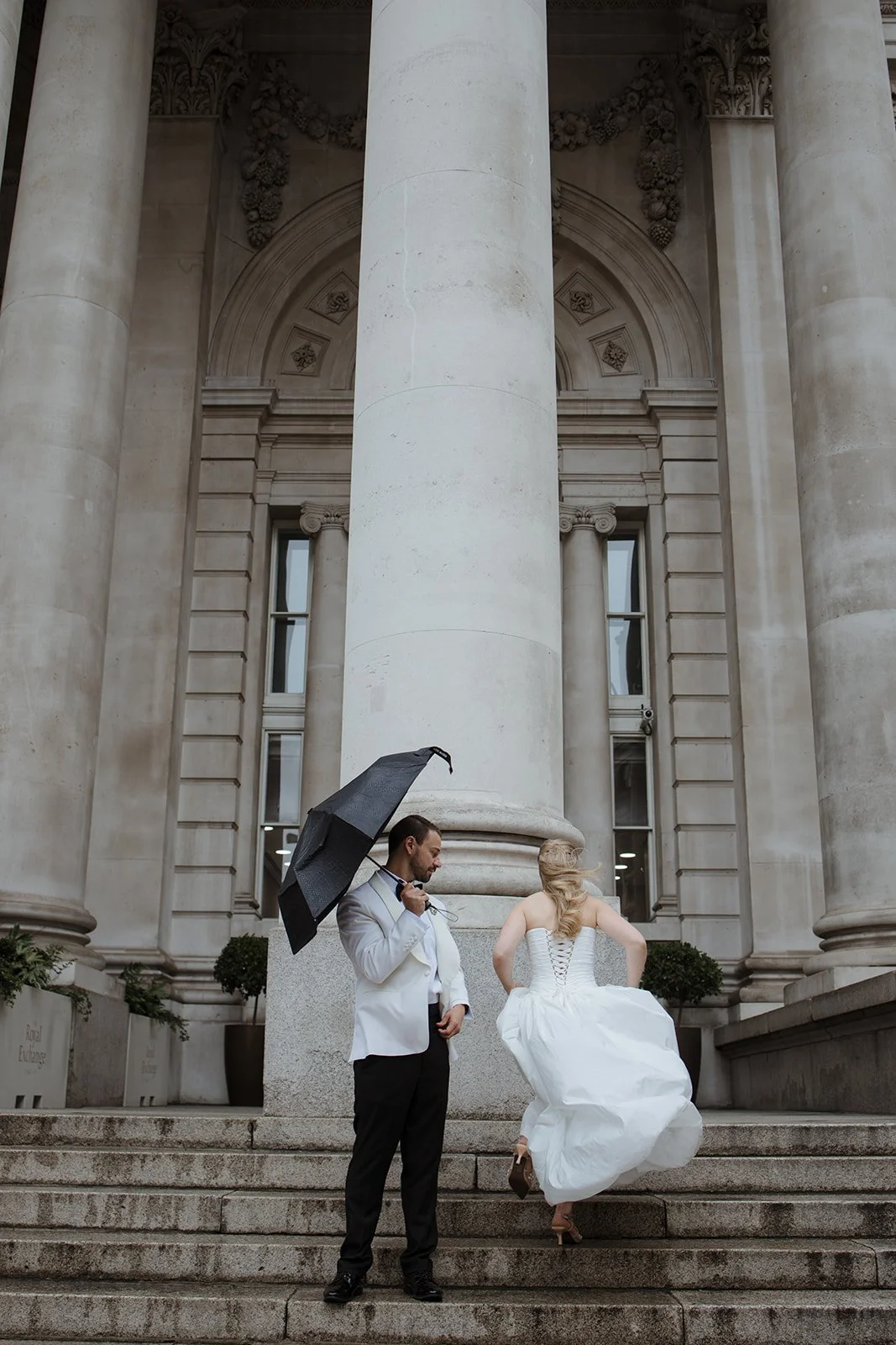 A couple dressed in wedding attire stands on steps in front of a large classical building with tall columns and ornate architectural details. The man holds a black umbrella and looks at the woman, who is lifting her wedding dress and has her back to 