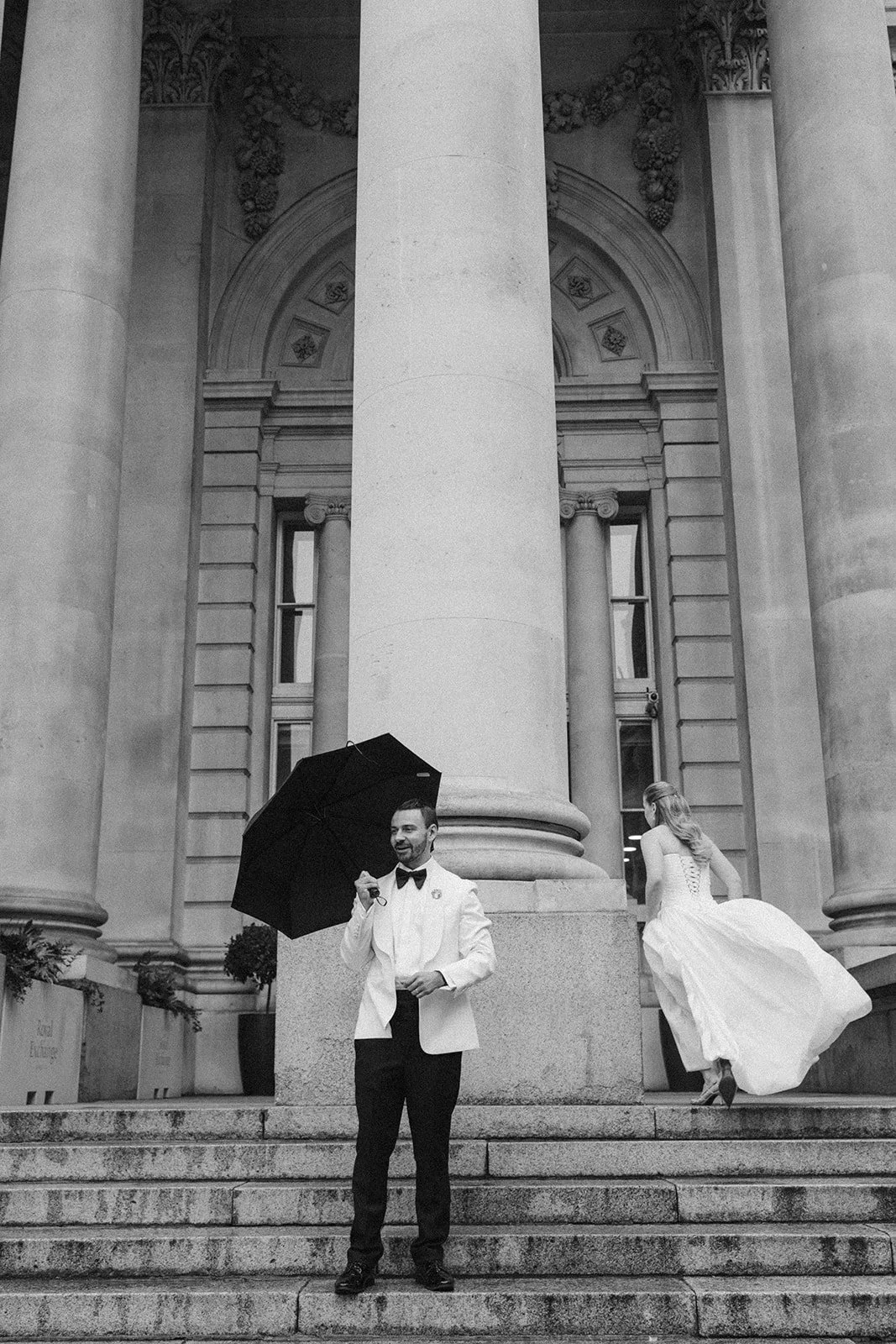 A black and white photo of a man in a tuxedo holding an umbrella, standing on steps in front of a large building with classical architecture. A woman in a wedding dress is ascending the steps, partially visible, with the back to the camera.