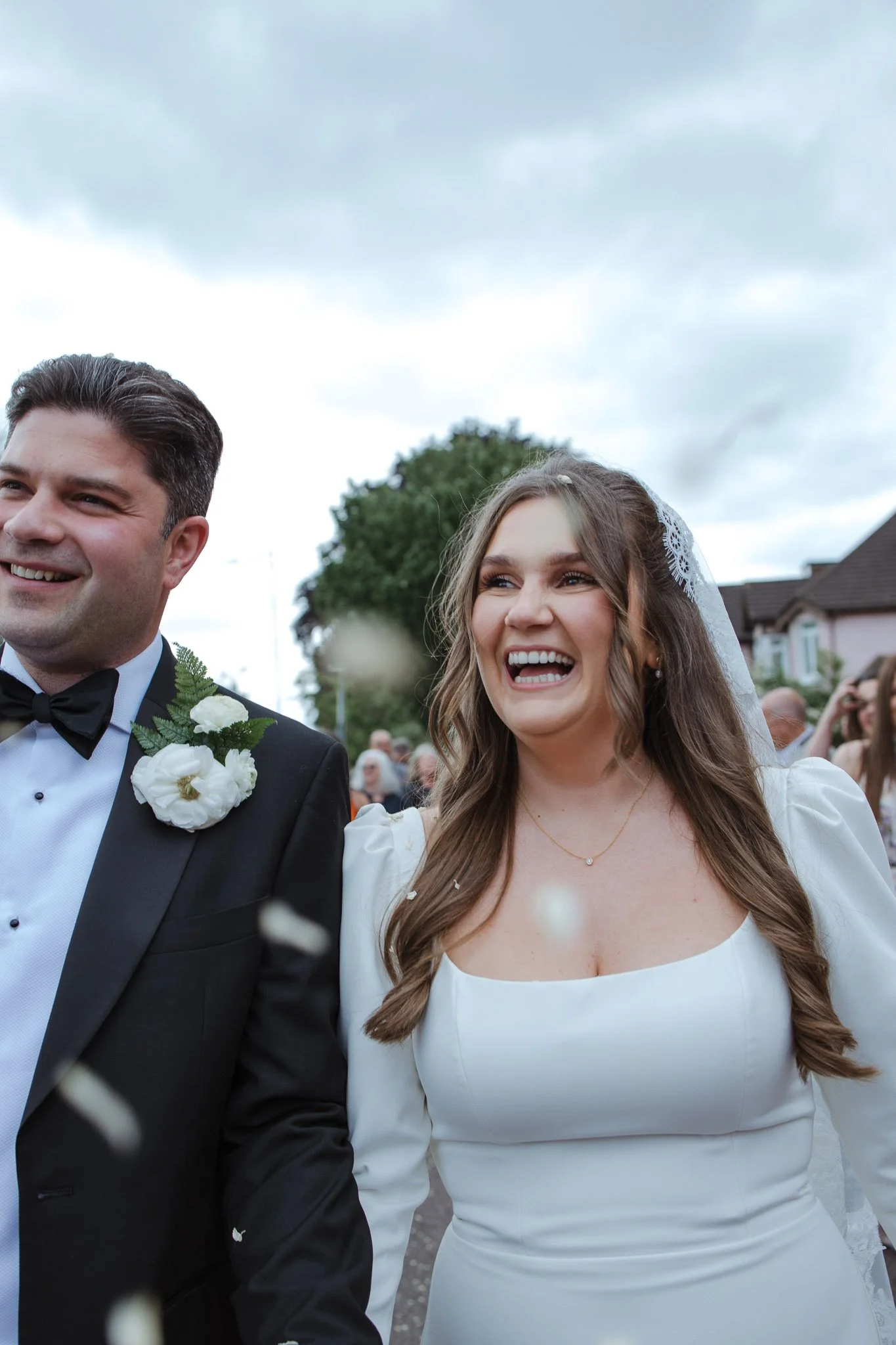 A smiling bride in a white wedding dress and a groom in a tuxedo at their wedding, outdoors during daytime with guests and houses visible in the background.