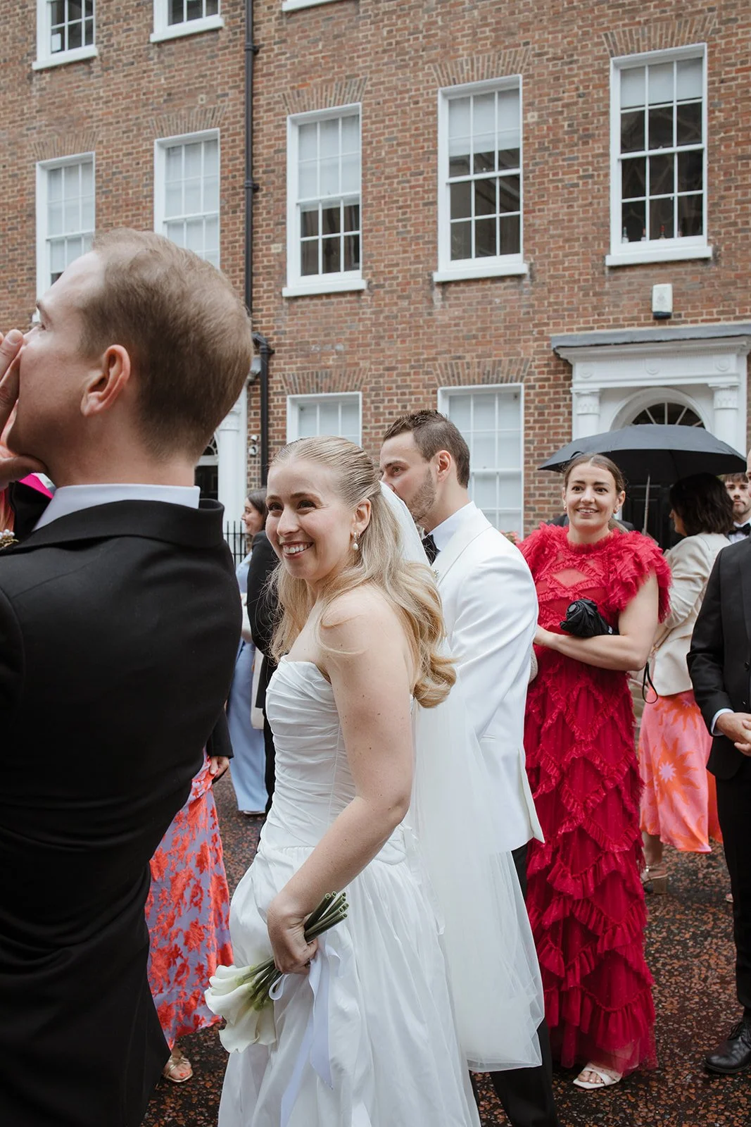 People dressed in wedding attire standing outside in front of a brick building, some smiling, one woman holding a small bouquet of white flowers, others holding umbrellas.