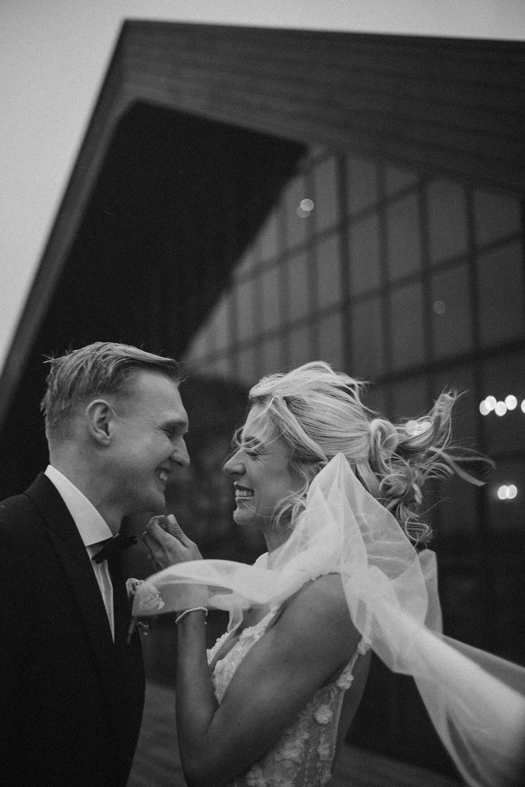 A black and white photo of a smiling couple, dressed in formal attire, close together, outside a modern building with large glass windows, sharing a joyful moment.