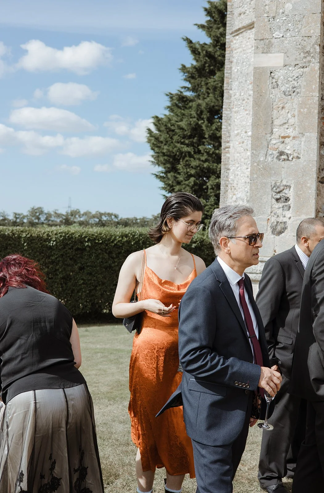 Group of people attending an outdoor event near a stone wall, with a young woman in an orange dress and several men in suits, under a partly cloudy sky.
