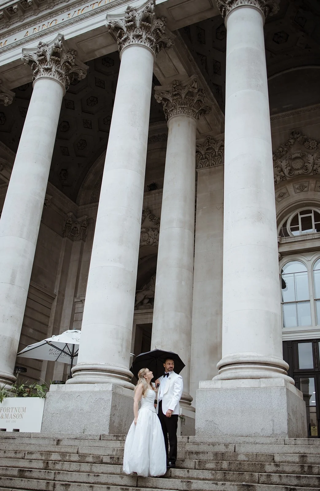 A bride and groom standing on stairs in front of a grand neoclassical building with large white columns, holding a black umbrella.