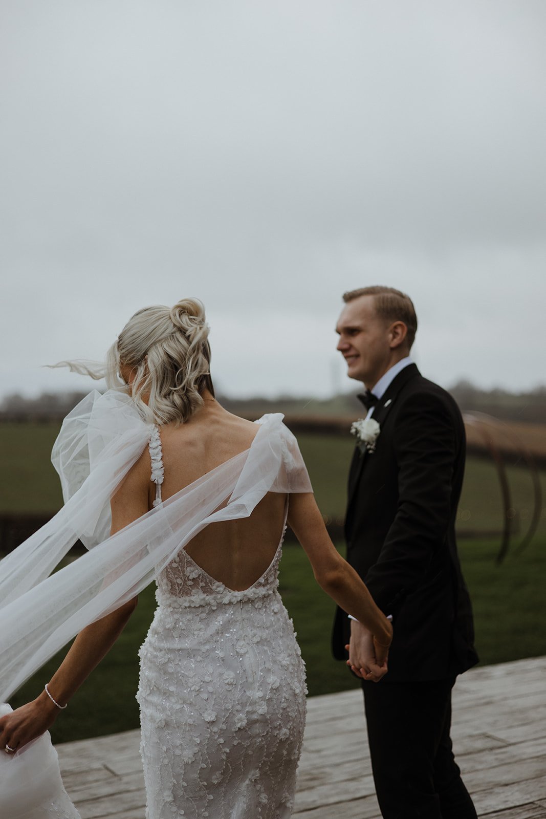 Bride and groom holding hands outdoors on a cloudy day, with the bride wearing a backless wedding dress with lace details and a flowing veil, and the groom in a black tuxedo.