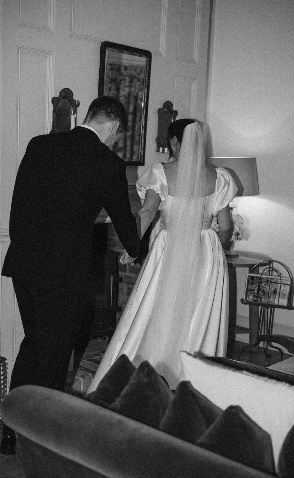 A bride in a wedding gown with a veil, and a groom in a suit, holding hands inside a home, preparing for a wedding.