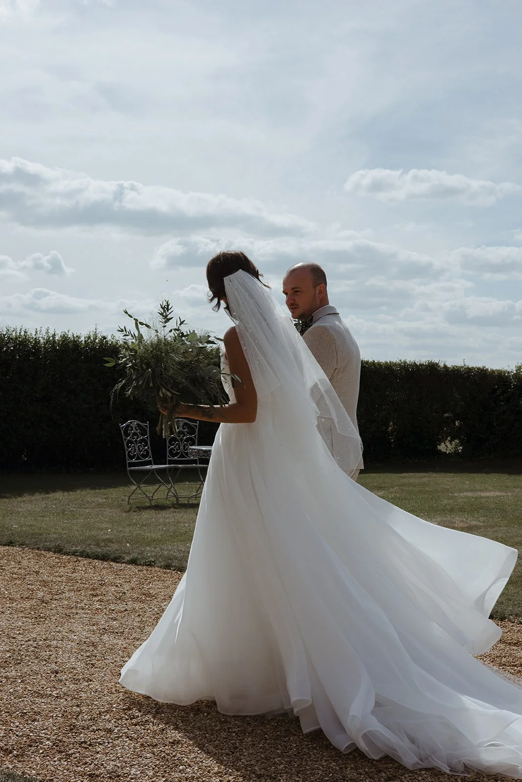 A bride in a white wedding gown holding a large bouquet standing next to a groom in a beige suit during an outdoor wedding under a cloudy sky.