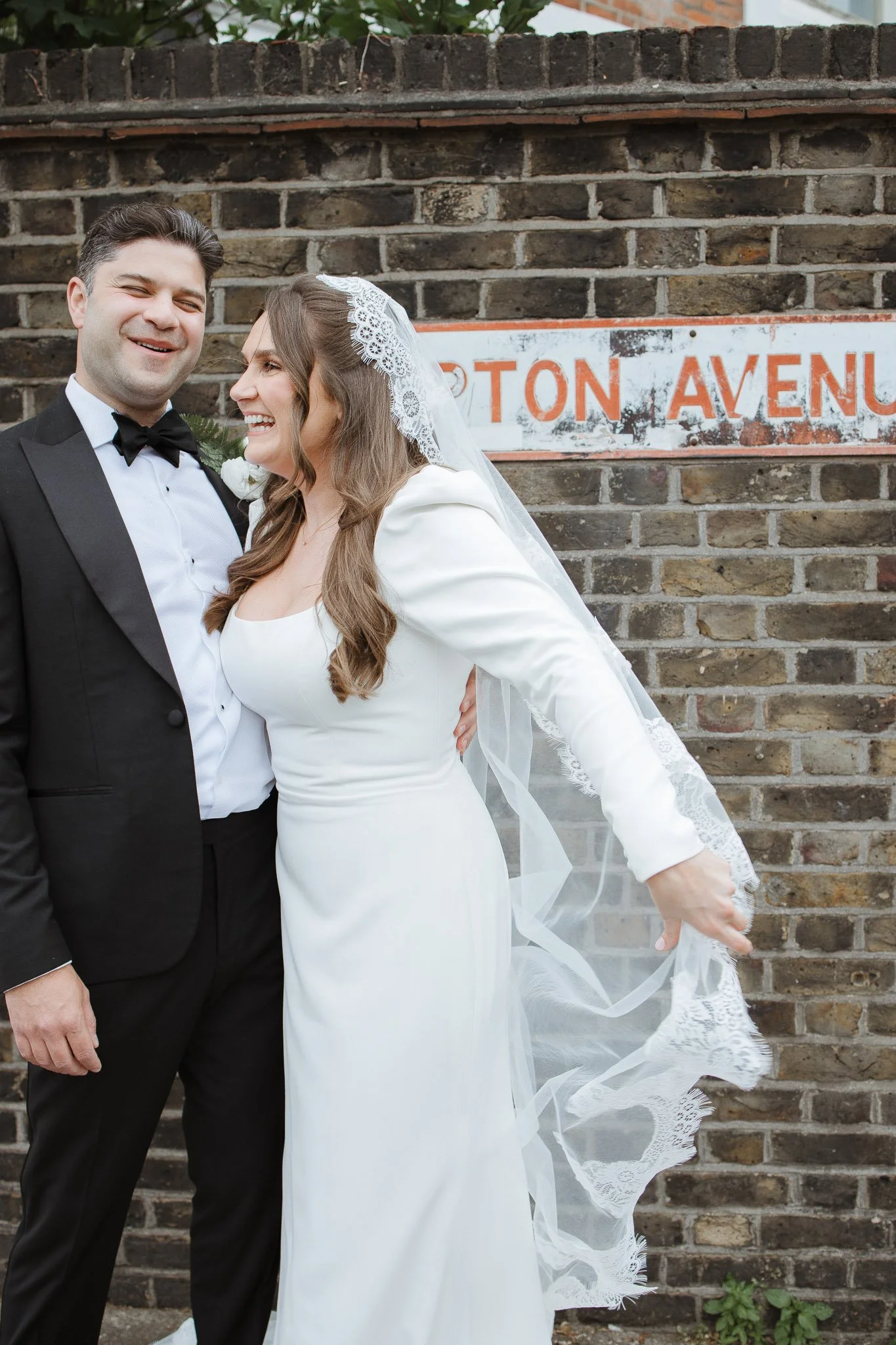 A newlywed couple in formal wedding attire, standing in front of a brick wall with a street sign that reads 'Baton Avenue'. The bride is wearing a white dress and veil, and the groom is in a black tuxedo with a bow tie. They are smiling and looking h