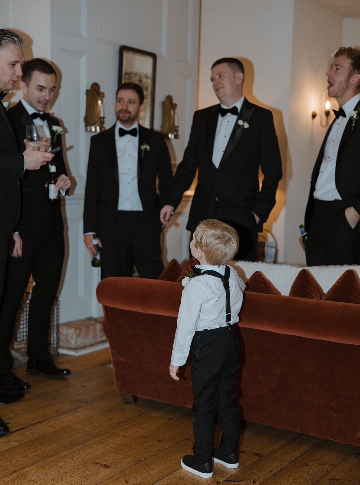 A young boy in black formal pants, white shirt, and suspenders stands in front of a vintage orange velvet couch, watching a group of men dressed in black tuxedos with bow ties, holding drinks, and socializing indoors.