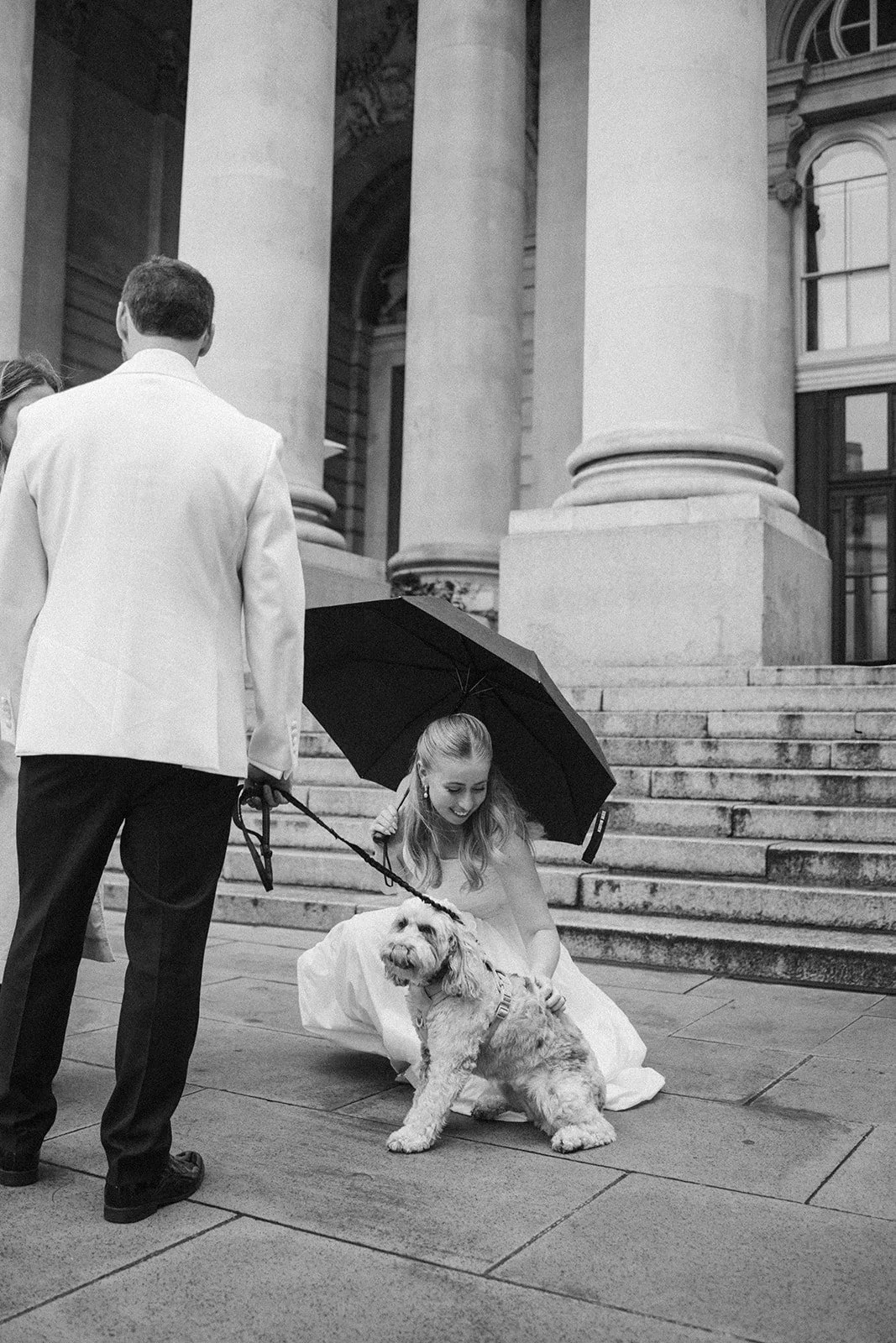 A woman in a wedding dress sitting on steps outdoors, holding a small dog, while a man in a tuxedo stands nearby. The woman is smiling and holding an umbrella over herself and the dog.