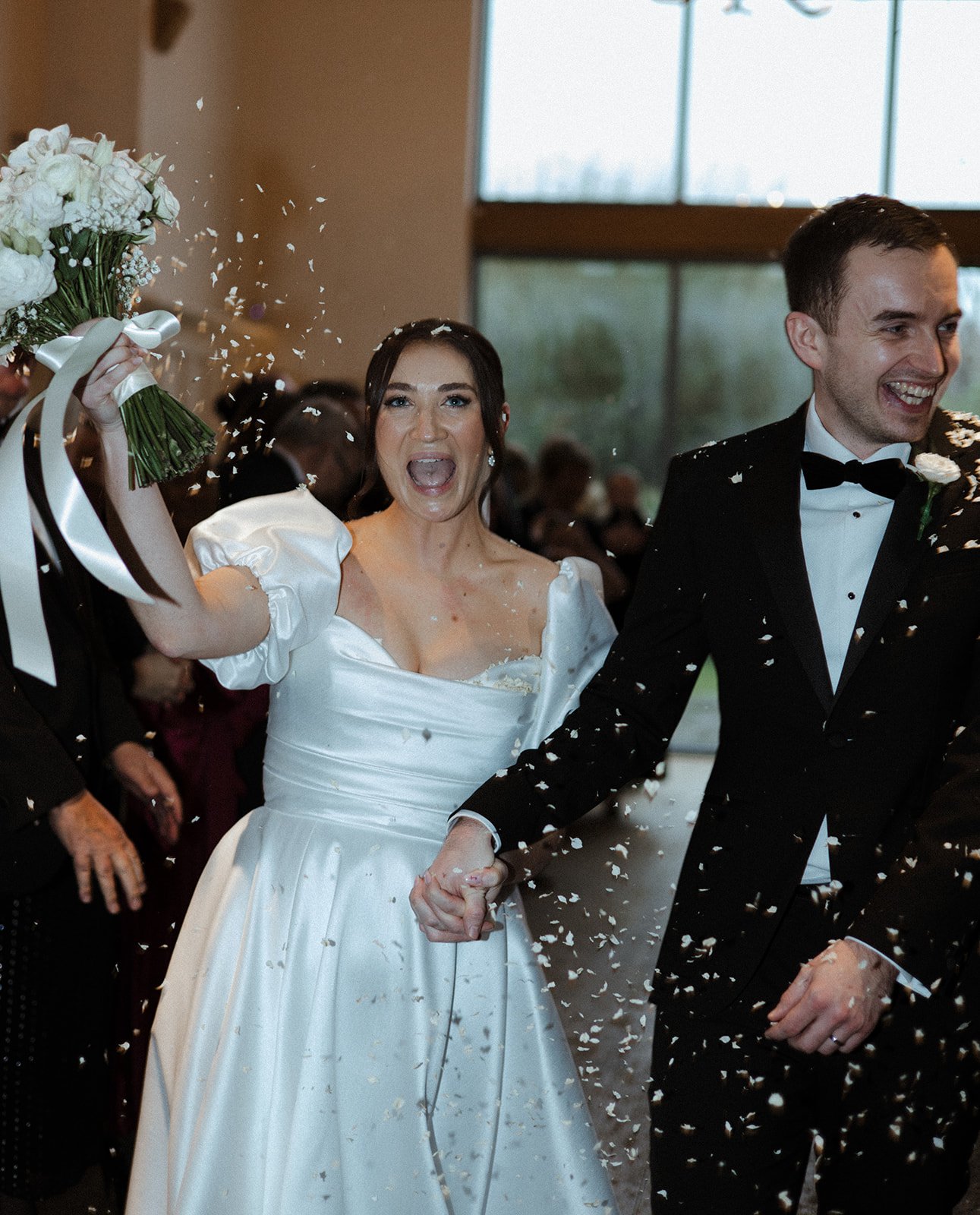 Newlywed couple celebrating their wedding with guests, holding hands, surrounded by falling confetti in a well-lit indoor venue.