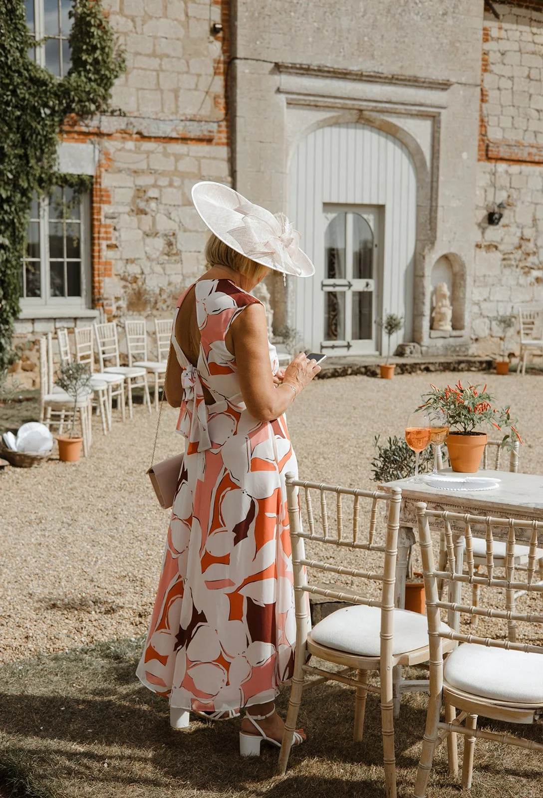 A woman in a floral dress and large hat stands outdoors using her phone at an event, with a stone building, chairs, and a table with a plant and glasses of rosé behind her.