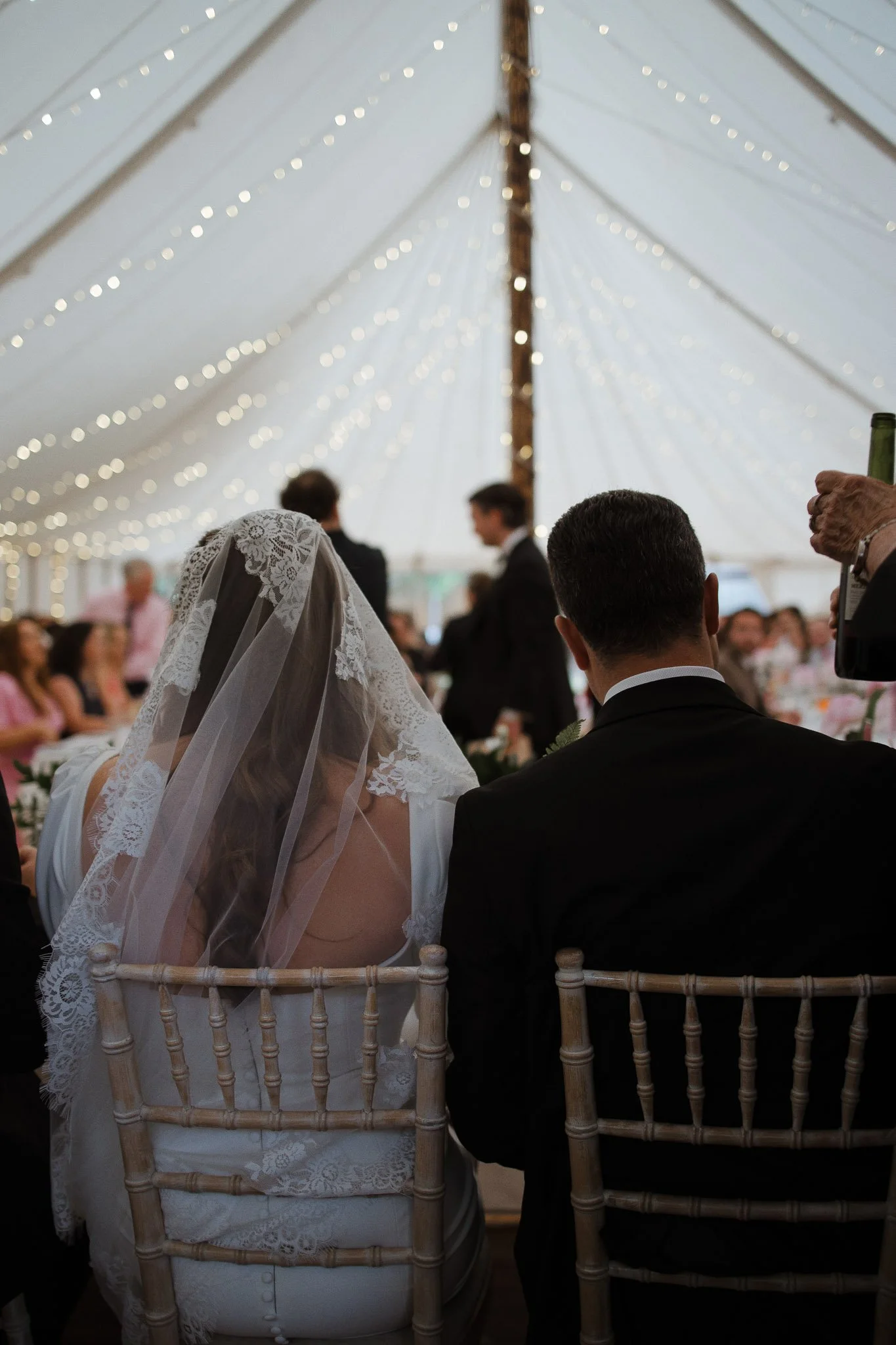 Wedding ceremony inside a decorated tent with string lights. A bride wearing a lace veil and white dress, and a groom in a black suit are seated.