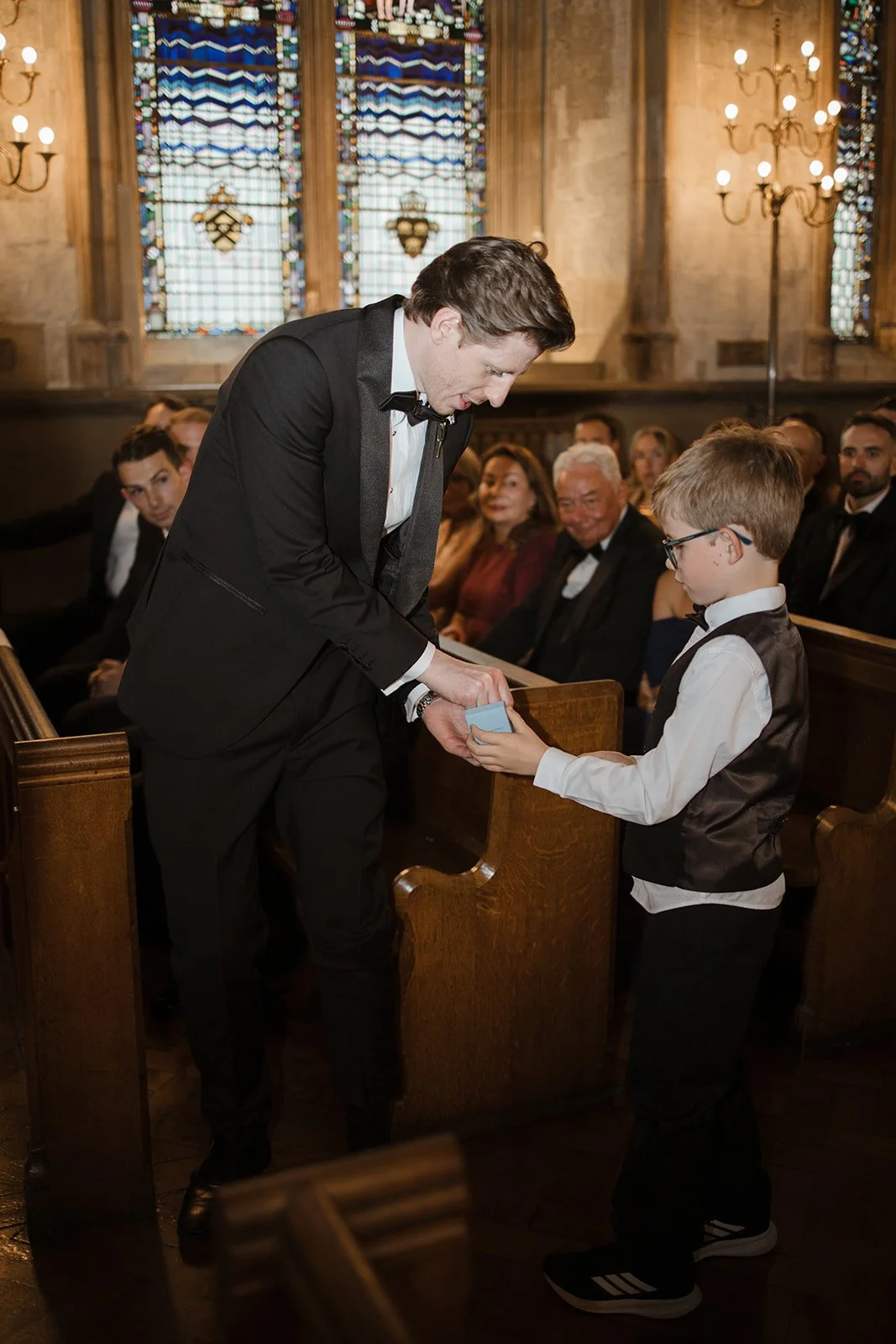 A man in a tuxedo giving an award or gift to a young boy in a vest and glasses during a ceremony inside a church with stained glass windows and chandeliers, with seated guests watching.
