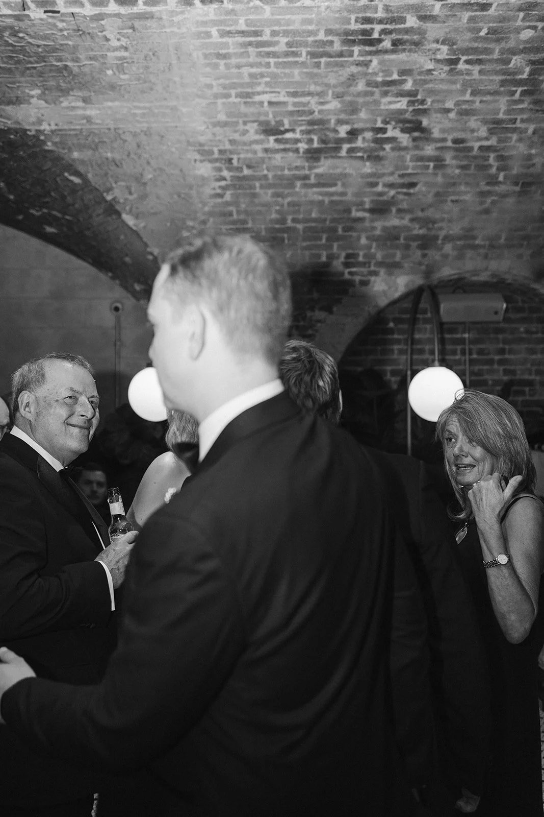 People socializing at a formal event in a room with arched brick ceilings, including men in tuxedos and a woman in a dress, some holding drinks.