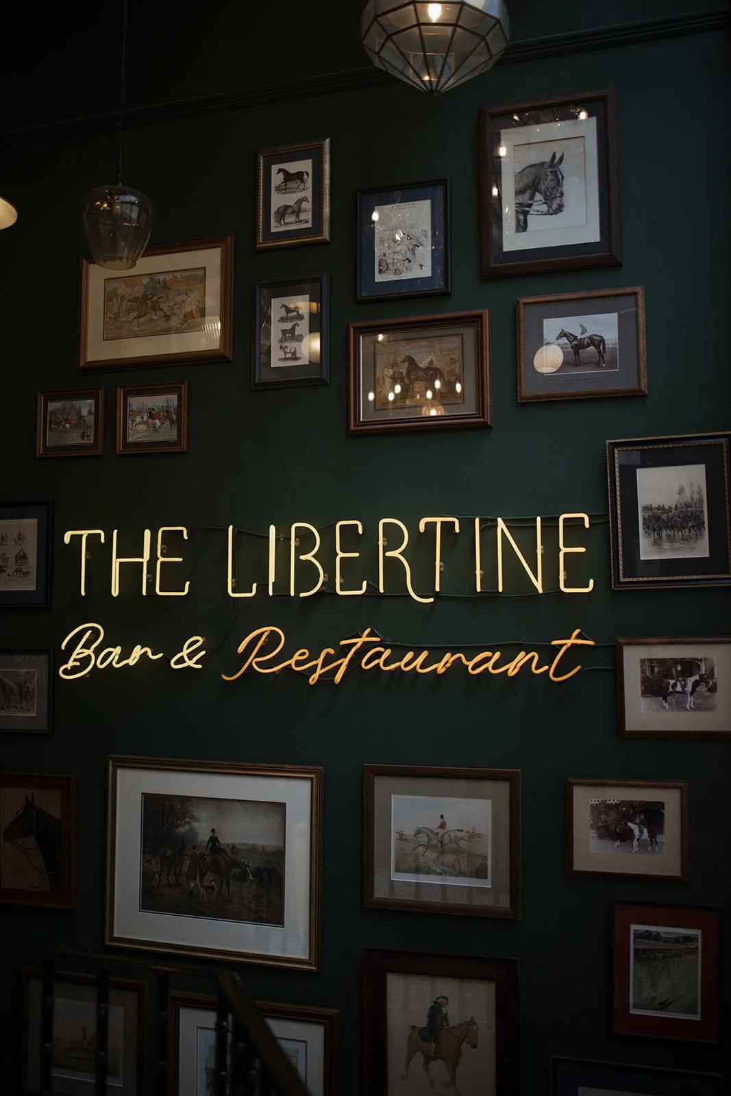 Interior of a bar and restaurant called The Libertine, featuring a green wall decorated with framed pictures of horses and a neon sign that says 'THE LIBERTINE Bar & Restaurant'.