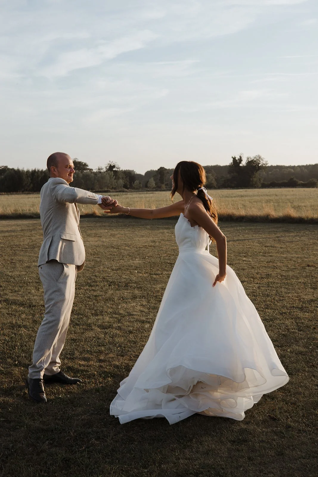 A bride and groom holding hands and dancing in an open field during sunset.
