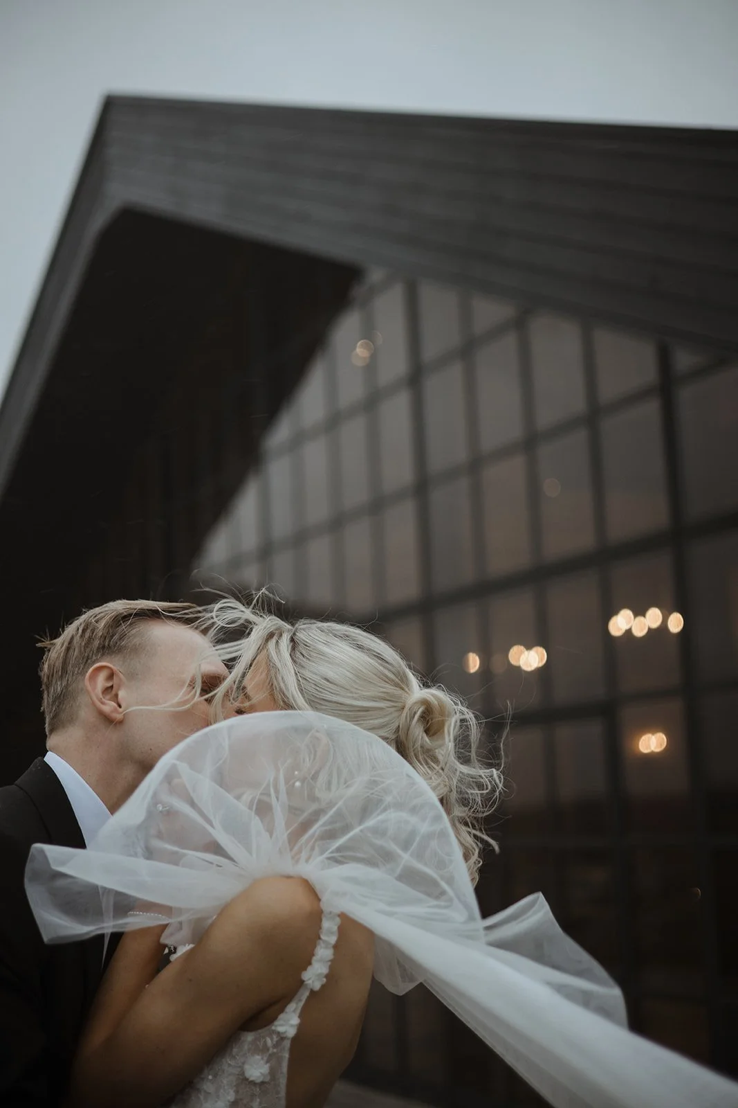 Couple sharing a kiss outside a modern building at dusk, woman in a wedding dress with veil, man in a suit.