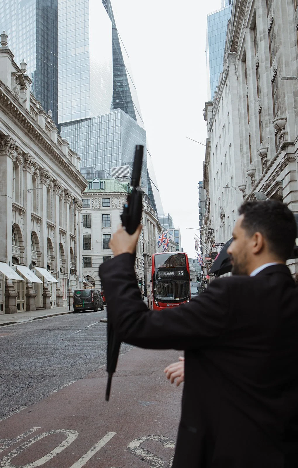 Man in a black suit holding an umbrella in a city street with buildings and a red double-decker bus behind him.