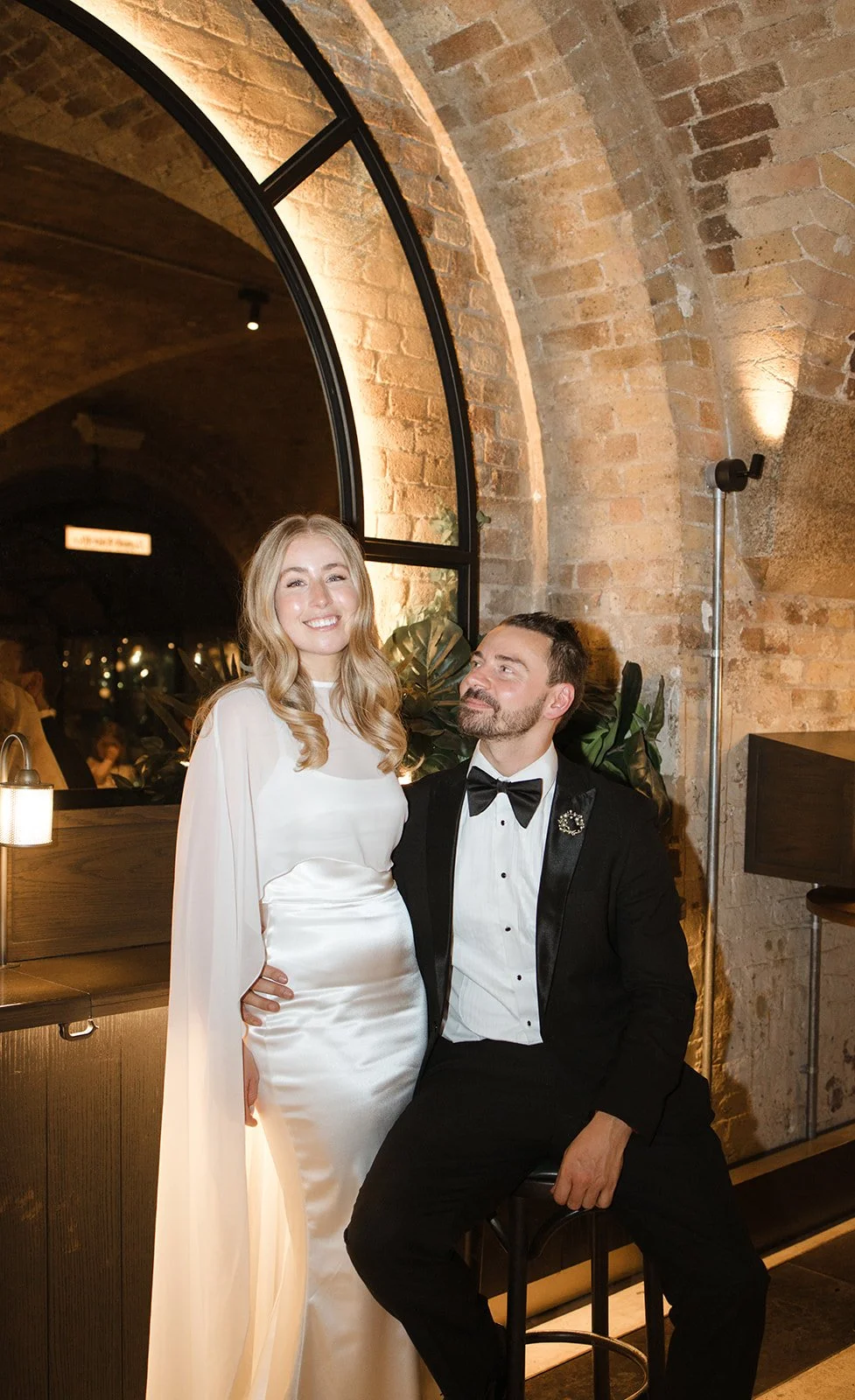 A couple dressed in formal attire sitting indoors near a large arched window with brick walls. The woman wears a white dress and the man wears a black tuxedo with a bow tie. The woman is smiling at the camera, and the man looks at her lovingly.