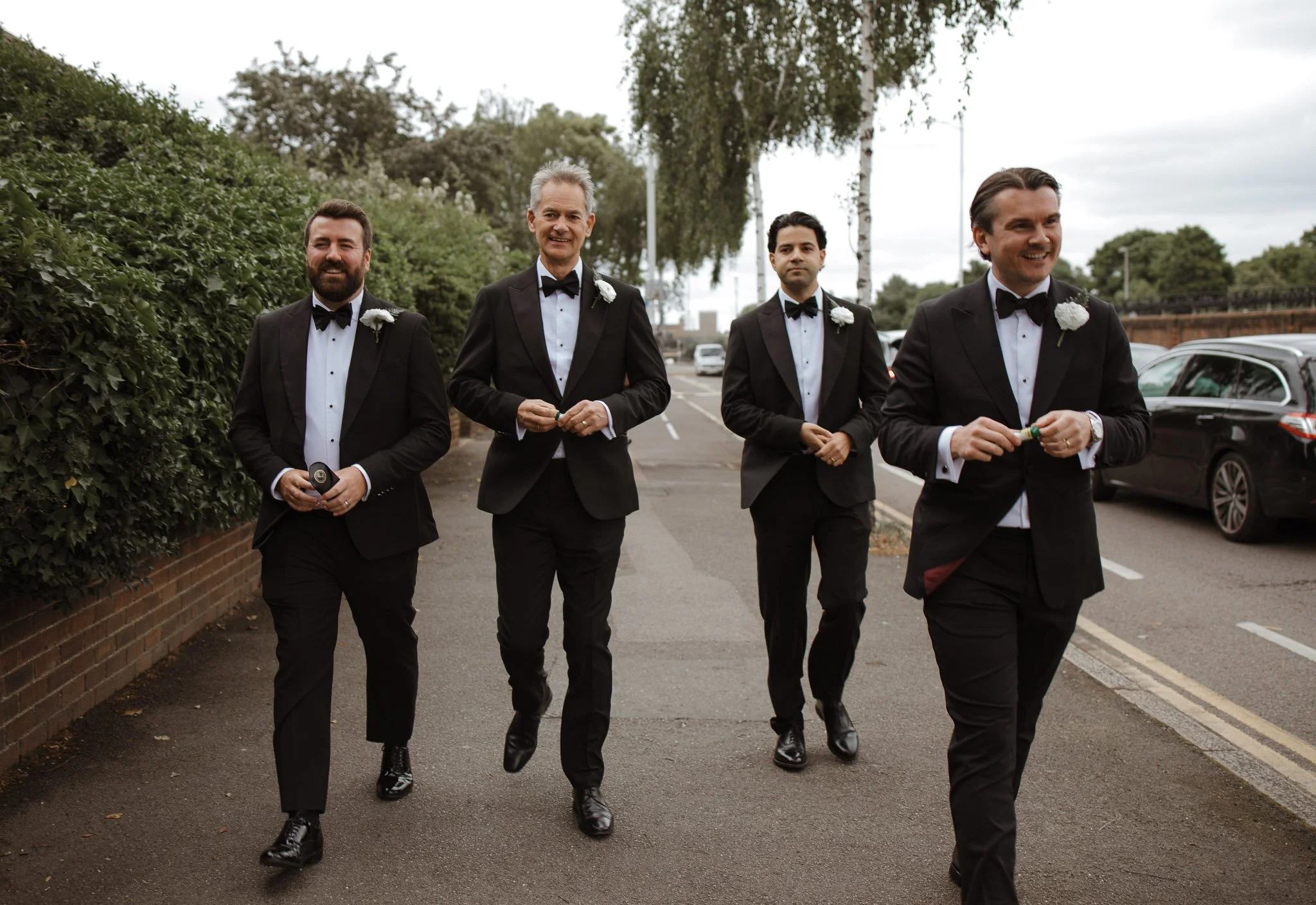 Four men in tuxedos walking on a sidewalk opposite parked cars and greenery, smiling.