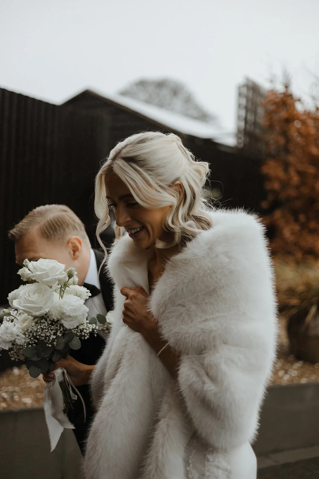 A woman with blonde hair smiling, wearing a white fur stole, holding her coat lapel, standing outdoors. A man in a tuxedo holding a bouquet of white flowers is next to her. The background includes trees with autumn-colored leaves and a dark building.