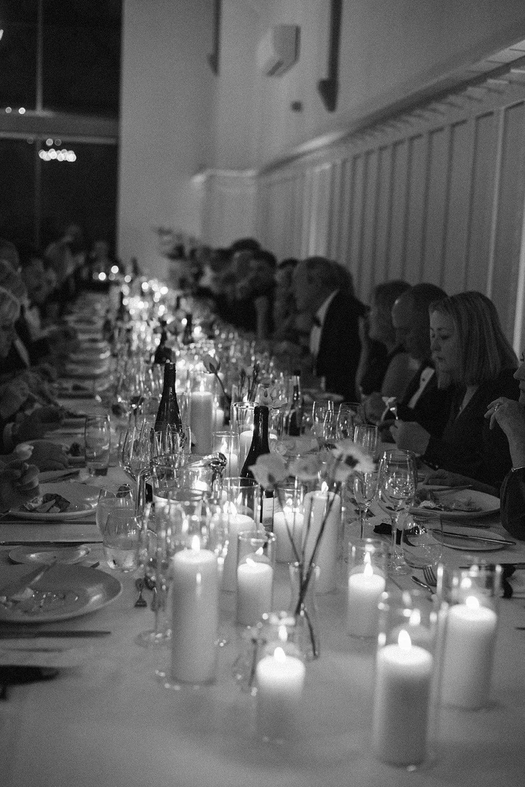 A black and white photo of a long banquet table set for a formal dinner, adorned with candles, wine glasses, and floral arrangements, with guests seated along both sides of the table.