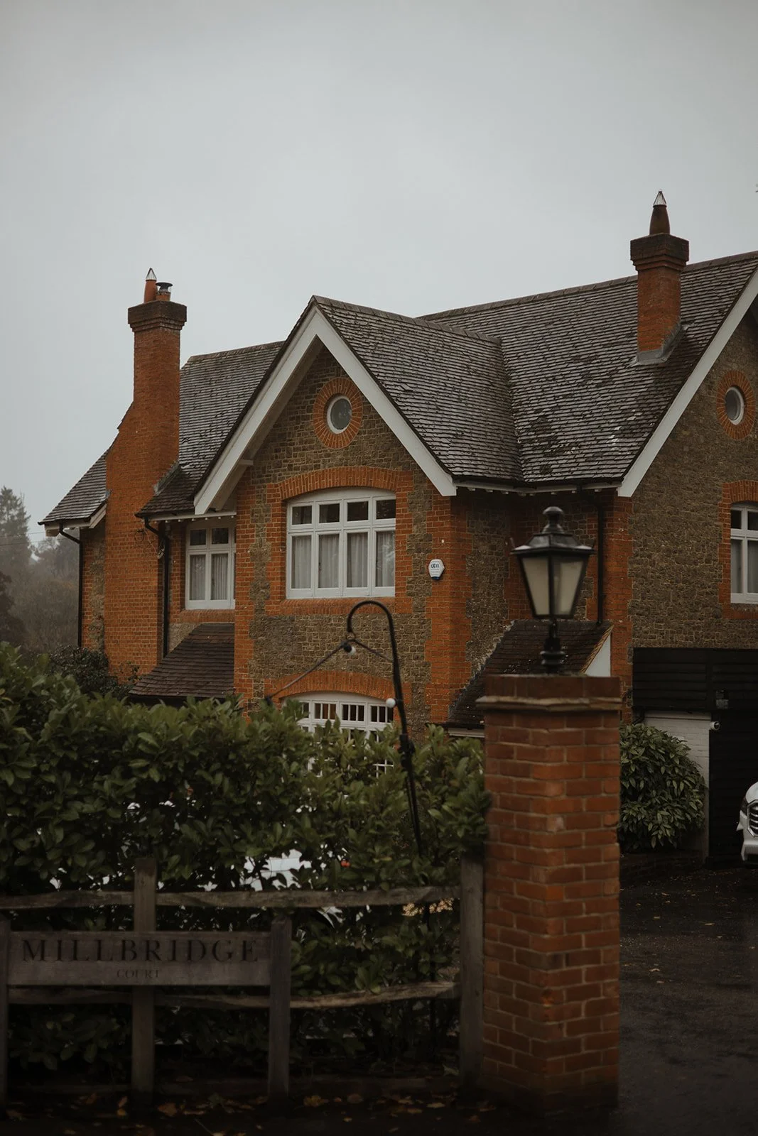 A large brick and stone house with multiple chimneys and gabled roof, surrounded by bushes, a brick pillar with a lantern, and a wooden fence with a sign that reads "Millbridge Court."