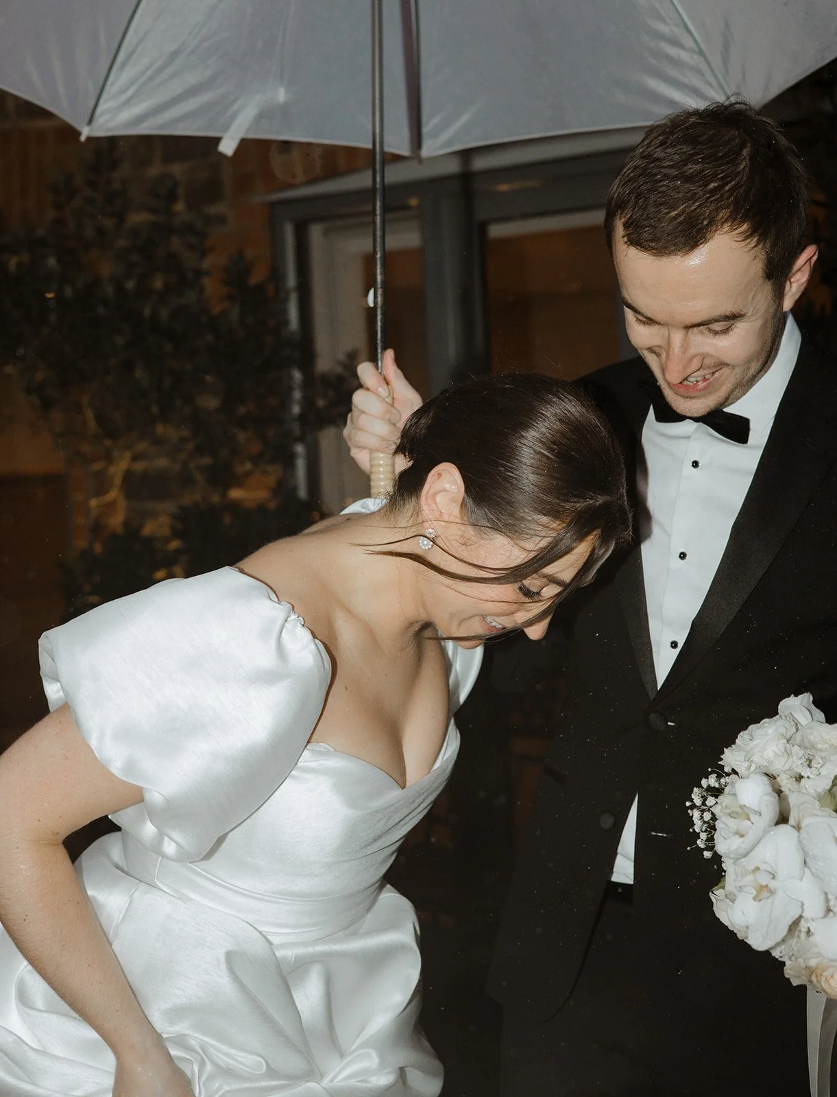 A bride and groom under an umbrella, smiling and looking down, celebrating their wedding.