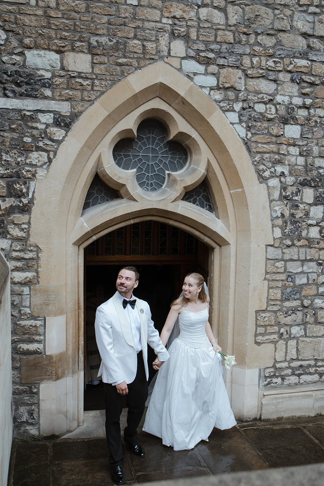 A bride and groom standing hand in hand outside a stone church, holding hands, with the bride in a white wedding dress and the groom in a white tuxedo jacket, black trousers, and bow tie, smiling and looking away.
