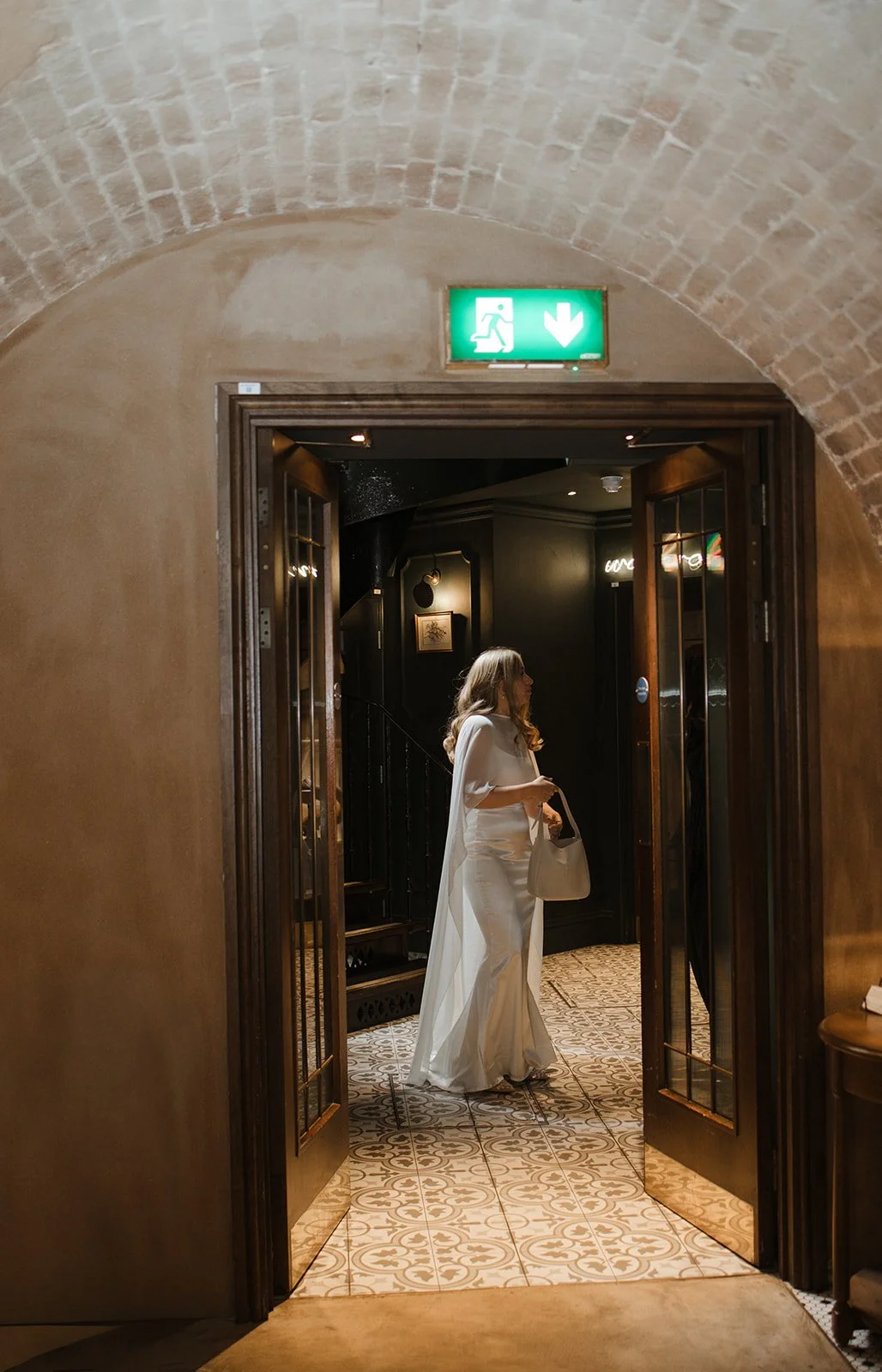 A woman in a white dress holding a bag, standing in an elegant, dimly lit interior near a doorway with a green emergency exit sign above.