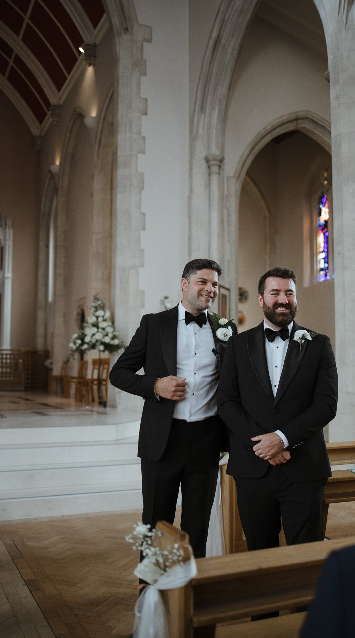 Two men dressed in black tuxedos with white shirts and black bow ties, each with a white flower boutonniere, standing inside a decorated church. The man on the right has a beard, while the man on the left has short dark hair, and both are smiling.