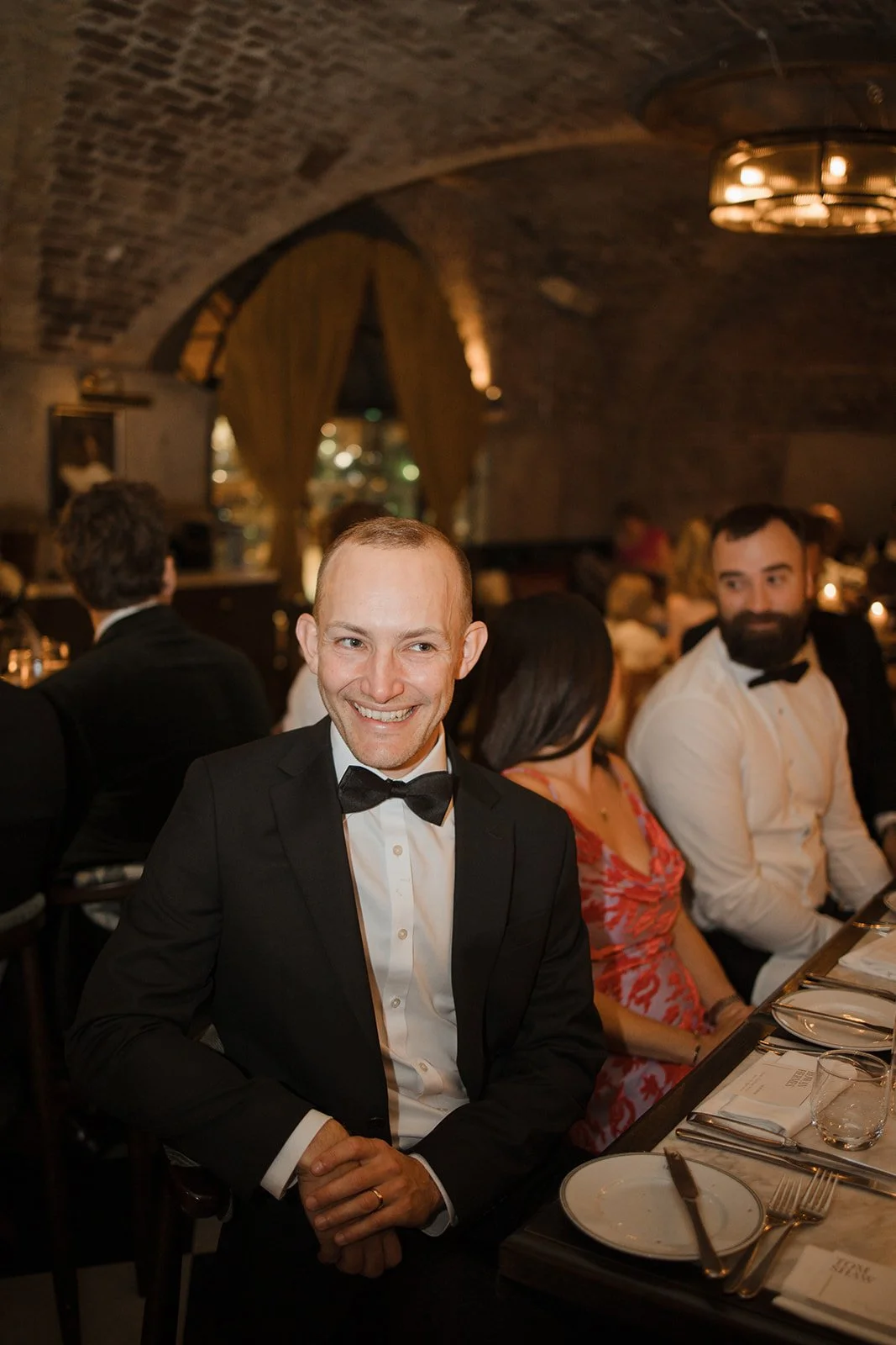 Man in a tuxedo smiling at a formal dinner event, seated at a table with plates, utensils, and glasses, with other guests in the background inside a decorated brick-ceiling venue.