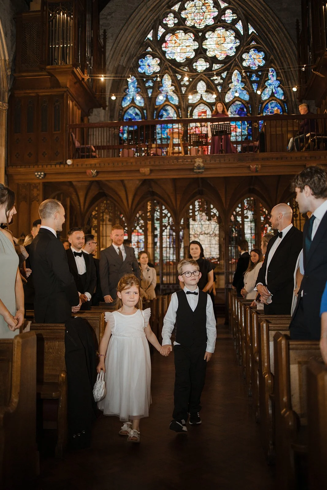 A young girl in a white dress and a young boy in a black vest and tie holding hands as they walk down the aisle of a church during a wedding ceremony. The church has tall stained glass windows and wooden pews, with other guests dressed in formal atti