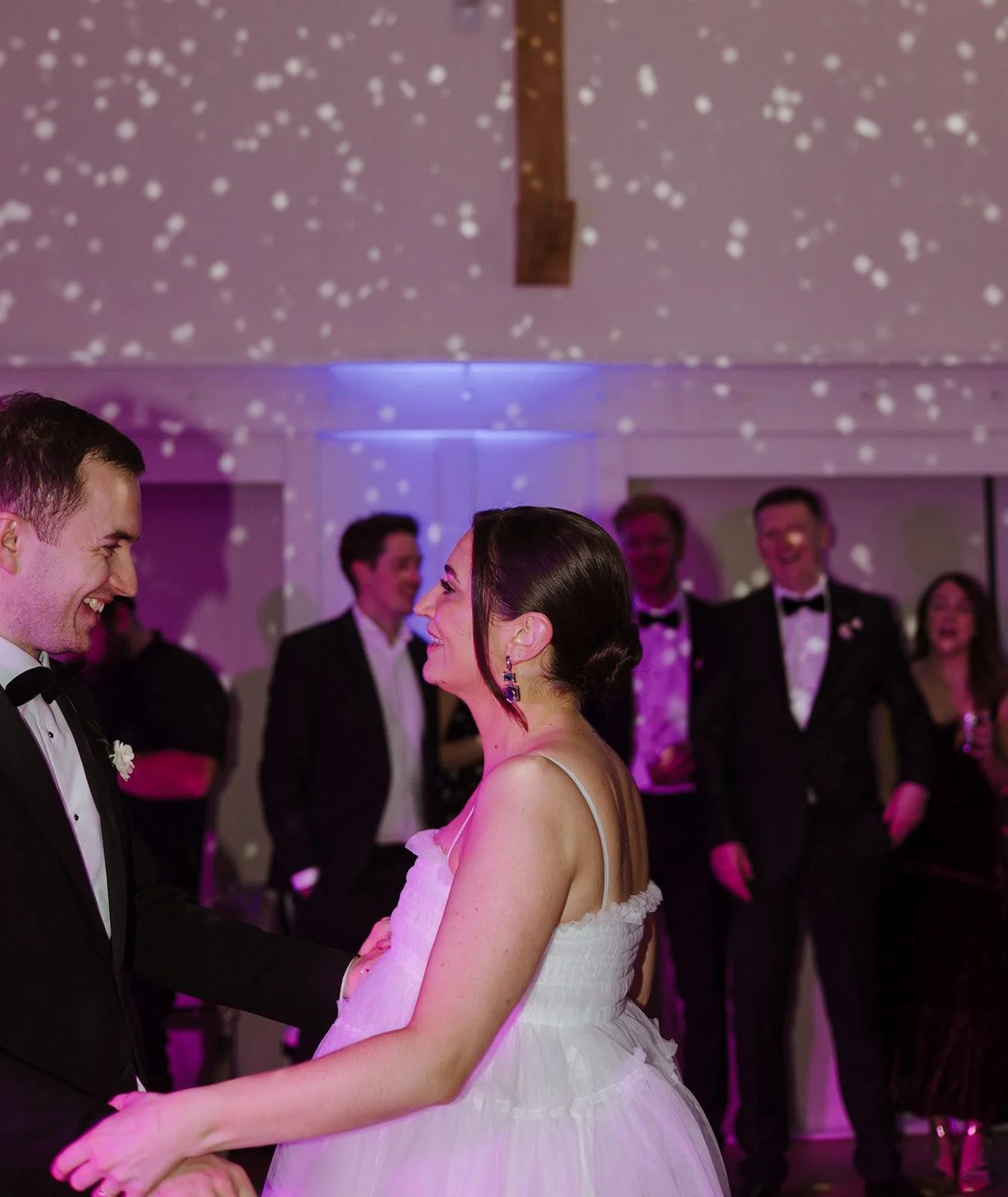 A bride and groom dance at their wedding reception, with friends in tuxedos and formal attire watching and smiling.