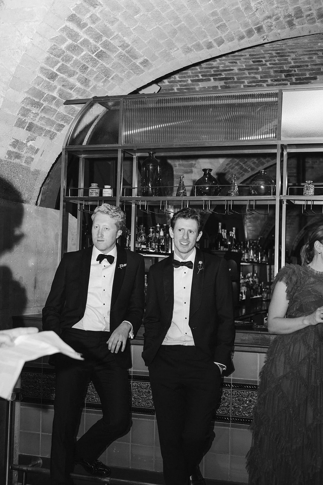 Black and white photo of two men in tuxedos with bow ties, standing and leaning against a bar in a brick-walled venue, with a woman partially visible on the right.