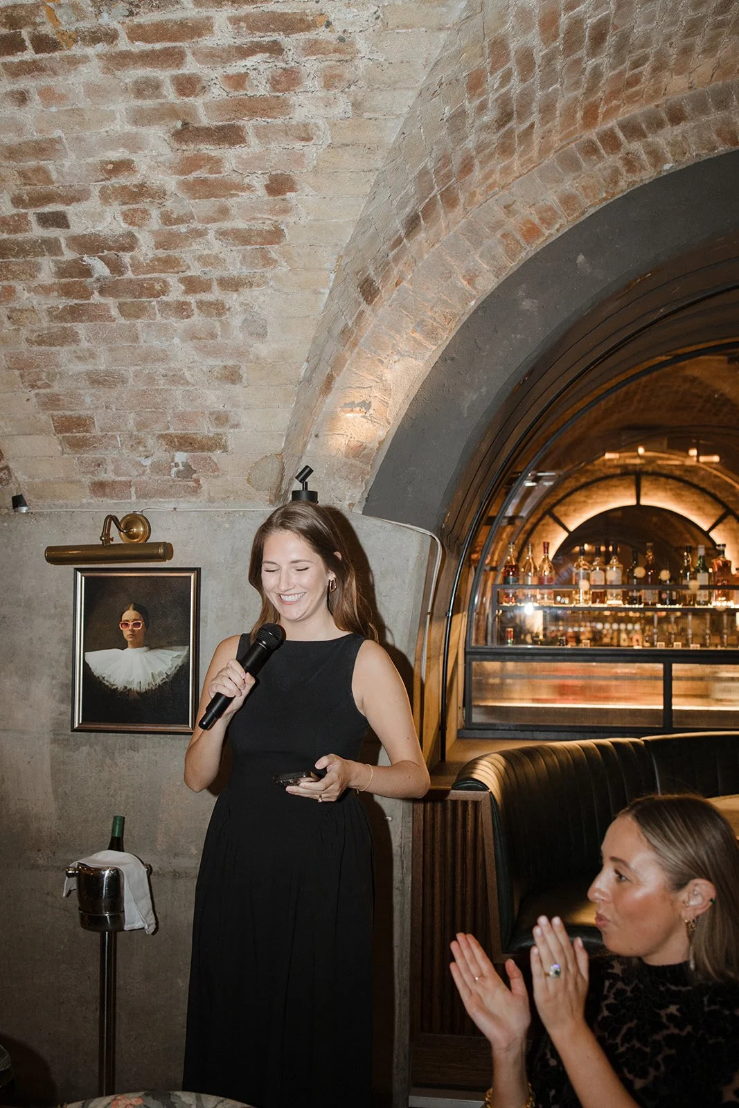 A woman in a black dress holding a microphone and smiling, standing in a restaurant or bar with exposed brick and a curved ceiling, while another woman claps and looks on.