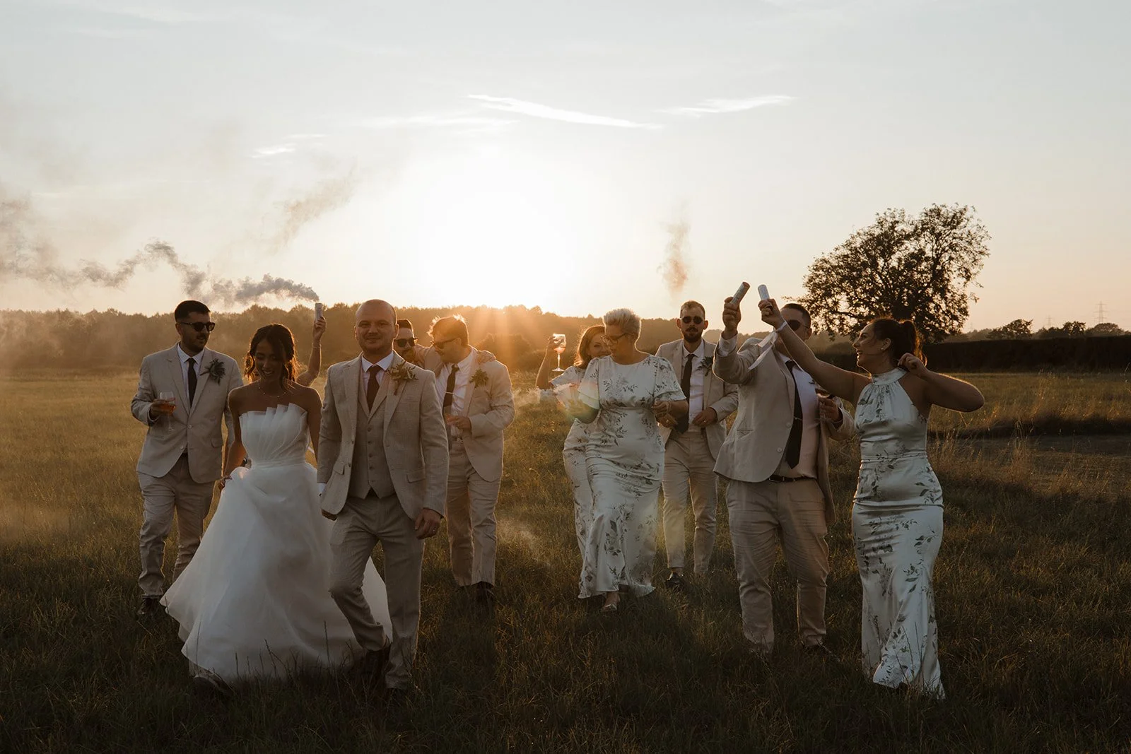 Group of people in formal attire, including a bride in a white wedding dress, walking outdoors during sunset in a grassy field, some holding drinks and taking photos.