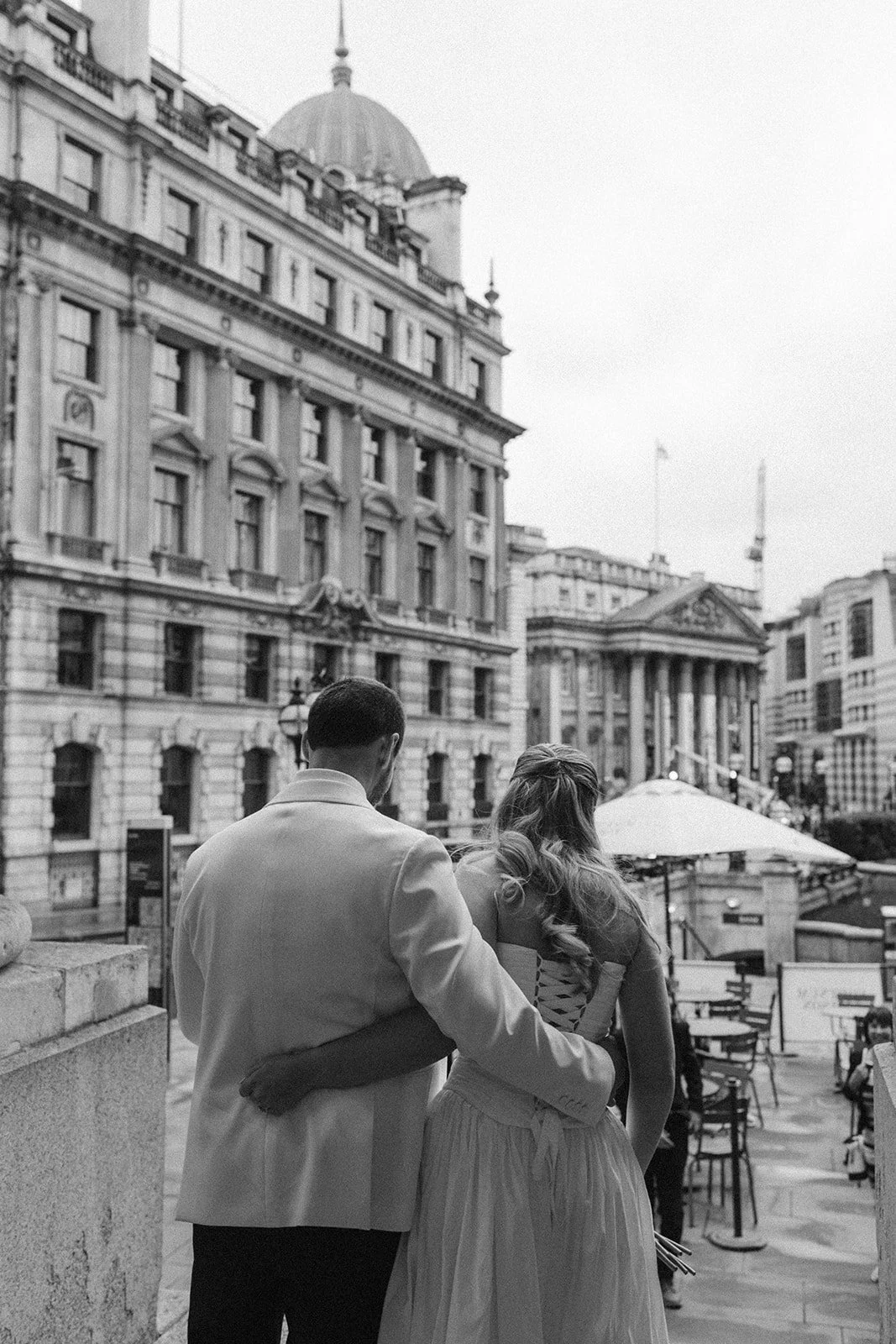 A black and white photo of a couple, with the man in a suit and the woman in a flowing dress, hugging on a city street with historic buildings and outdoor seating.