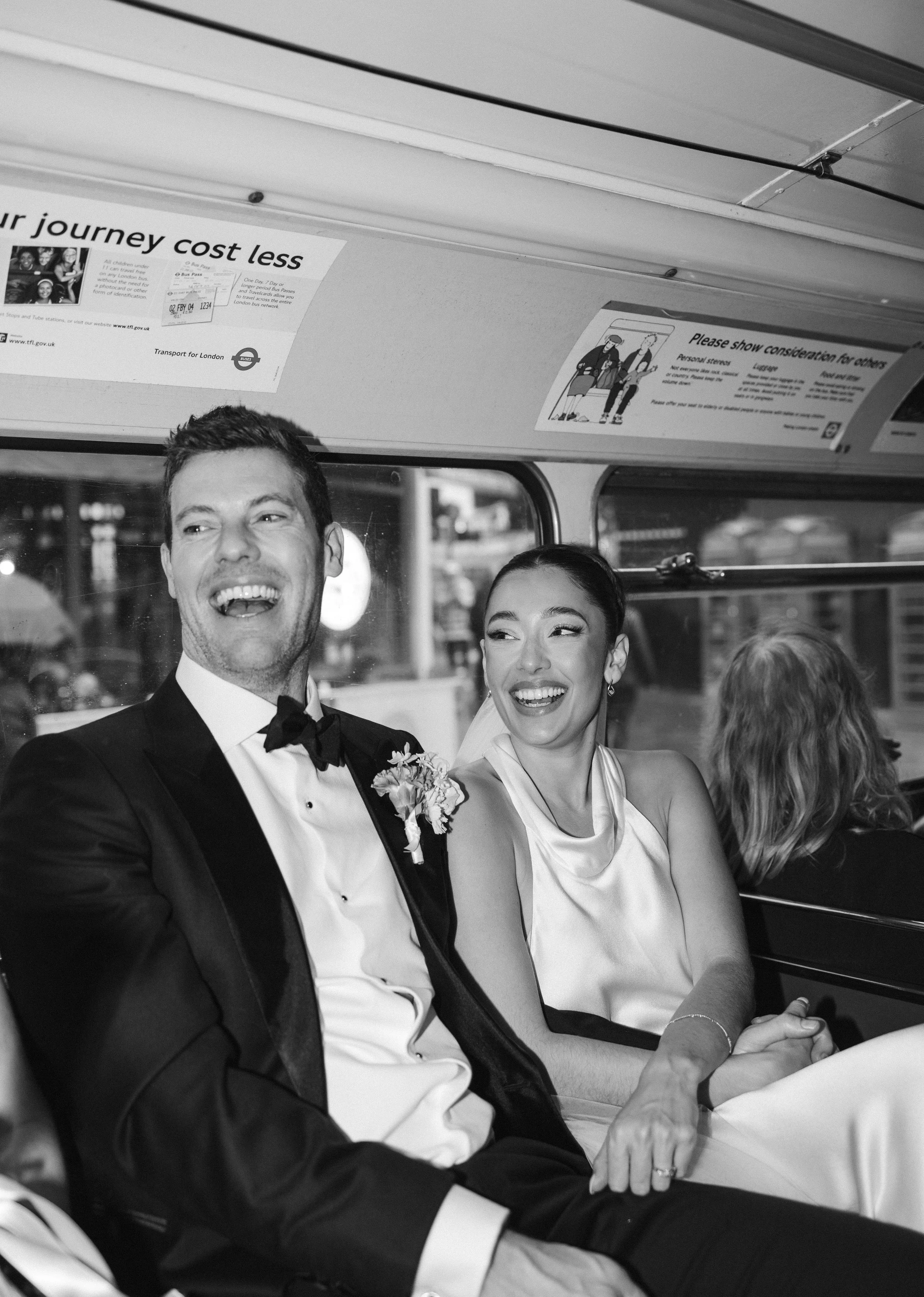 A black and white photo of a happy couple dressed in formal attire, sitting in a vehicle with several other passengers. The man is wearing a tuxedo and bow tie, and the woman is in an elegant dress. They are holding hands and smiling, with the woman's partner visible next to her.