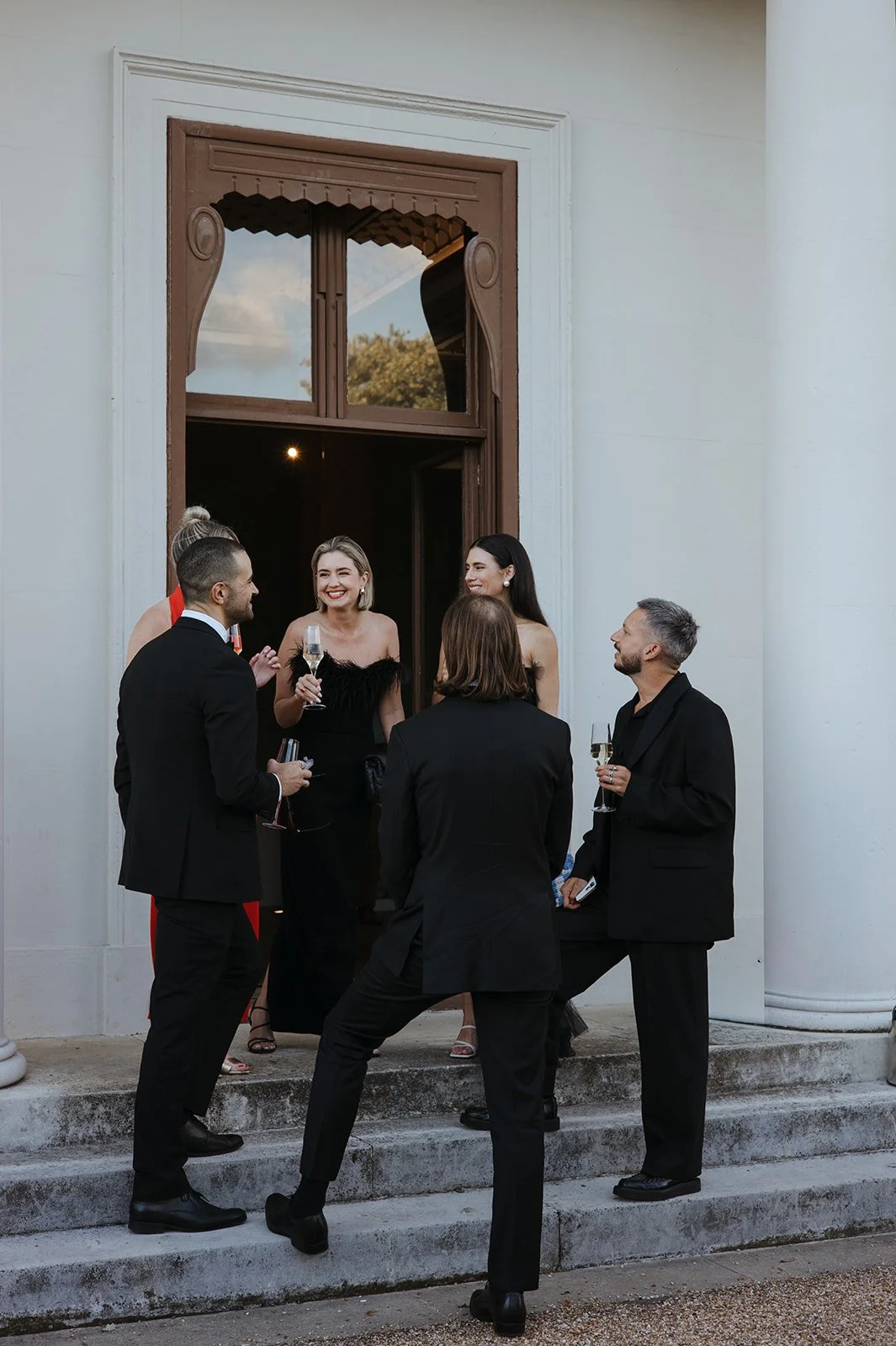 Group of people dressed in formal attire standing and socializing outside a building with steps, holding glasses of champagne, smiling and engaging in conversation.