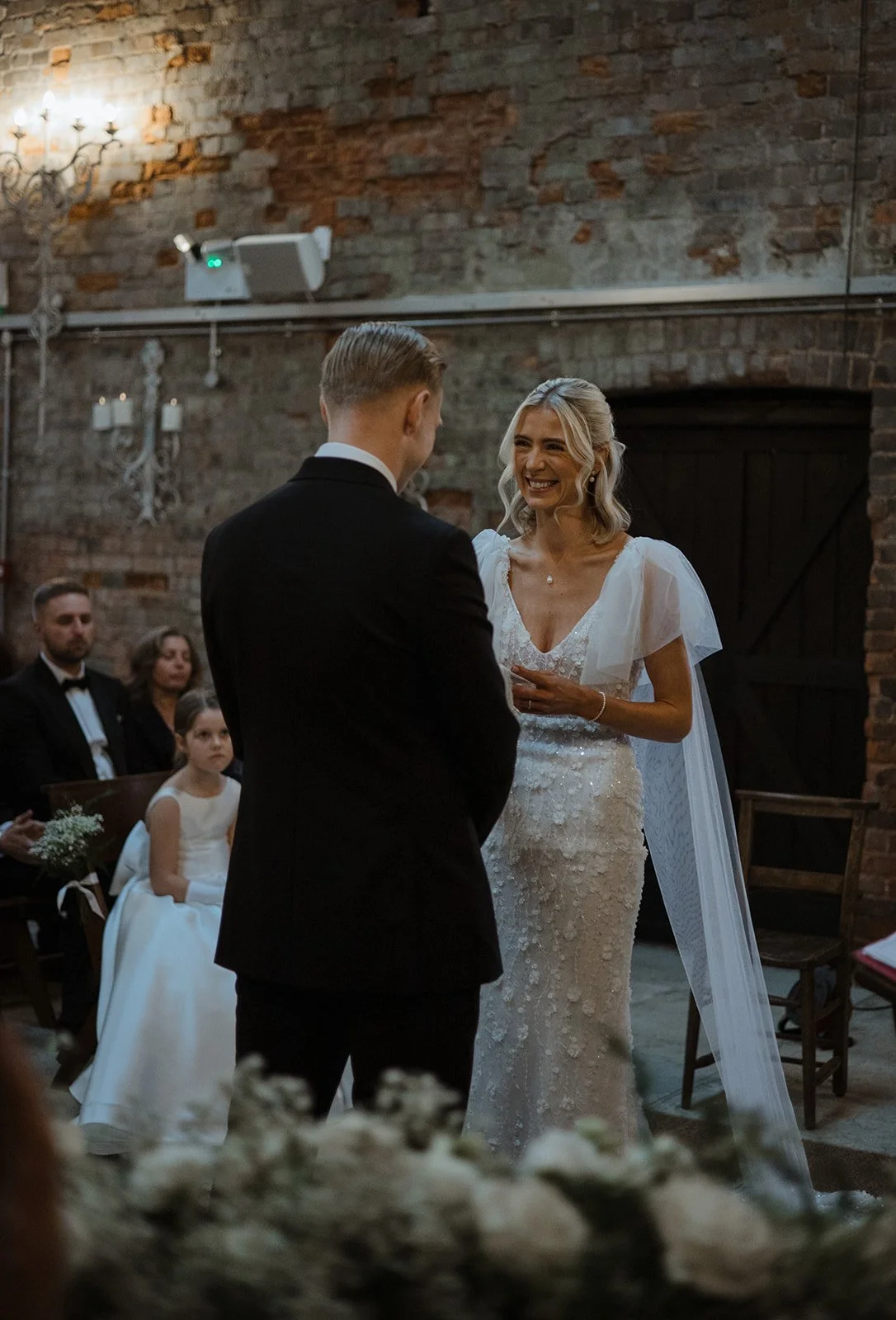 Bride and groom exchanging vows during wedding ceremony in rustic venue with exposed brick wall, chandelier, and seated guests.