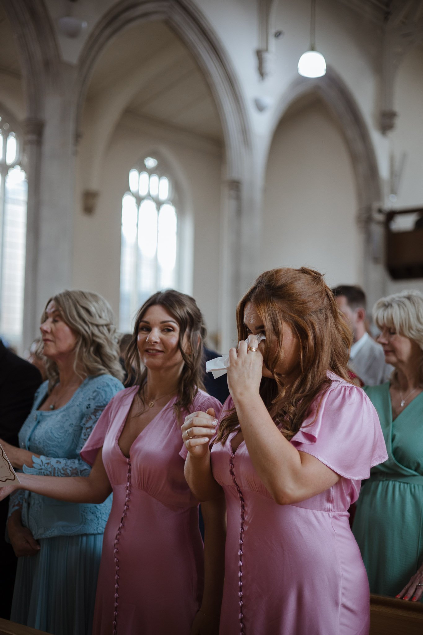 A group of women dressed in colorful dresses, standing inside a church during a wedding ceremony. One woman is crying and wiping her eyes, while others are looking on.
