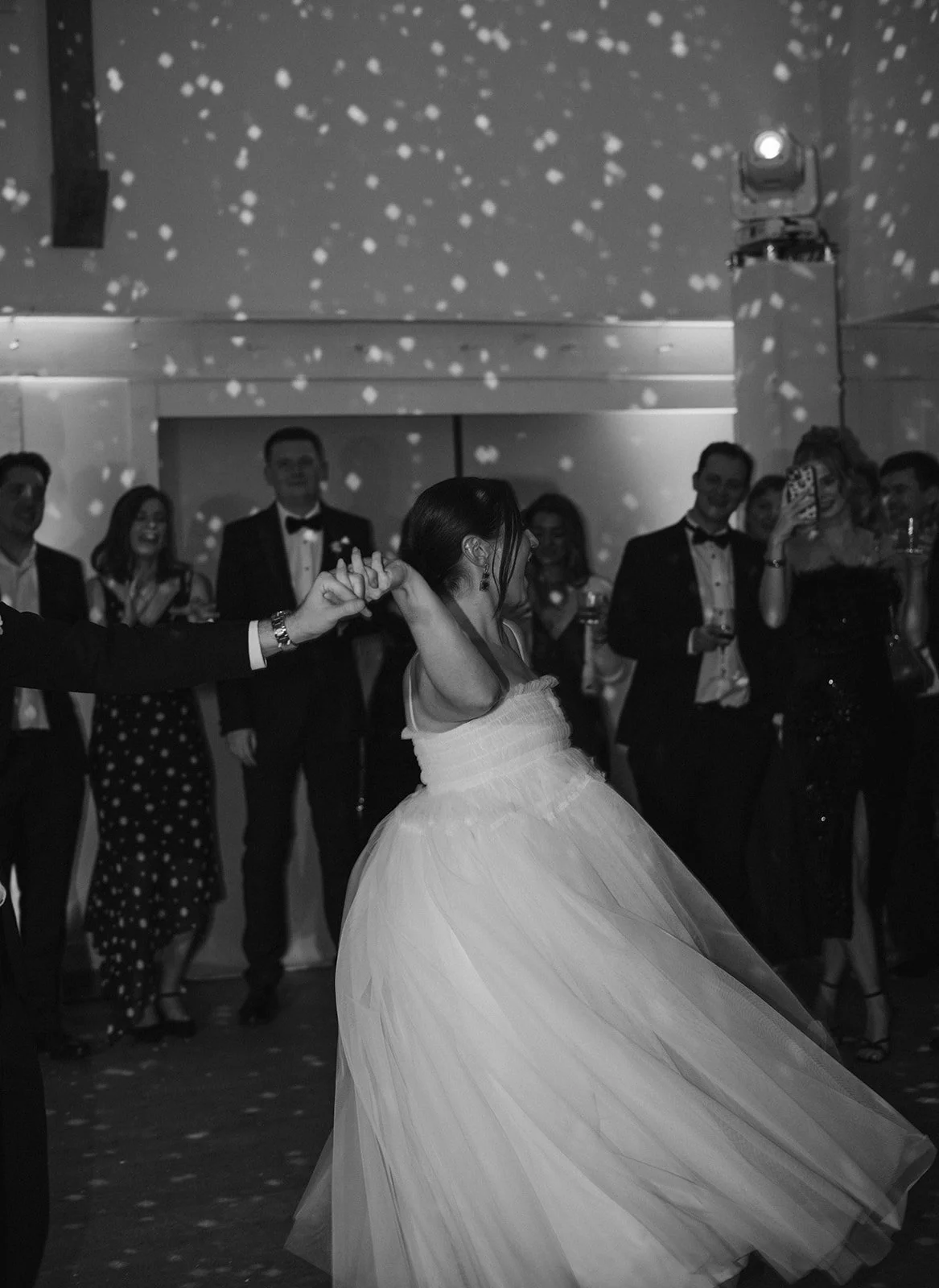 Black and white photo of a woman in a wedding gown dancing at her wedding reception surrounded by guests in formal attire.