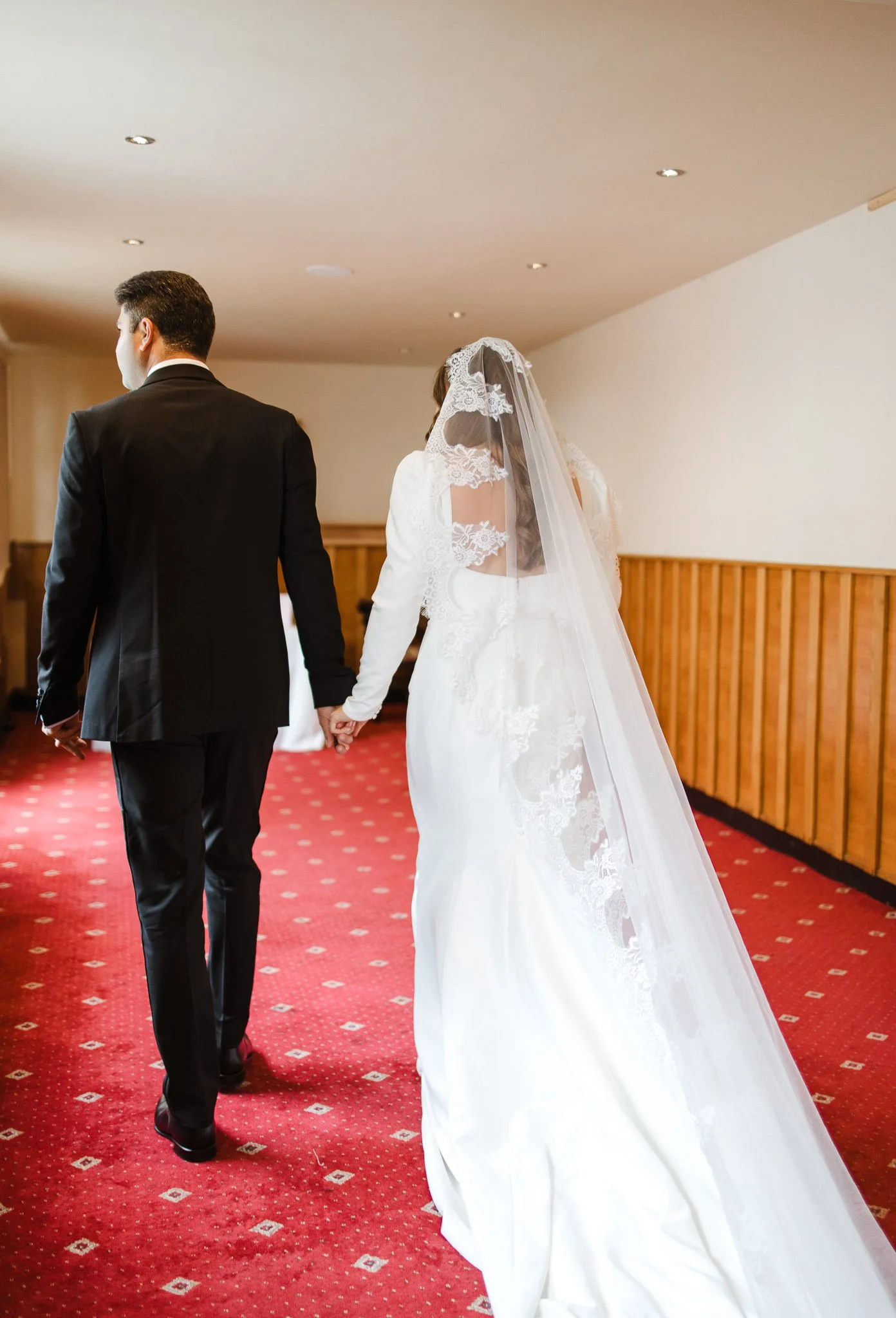 A bride and groom holding hands, walking away, with the bride wearing a white wedding dress and veil and the groom dressed in a black suit, in an indoor setting.