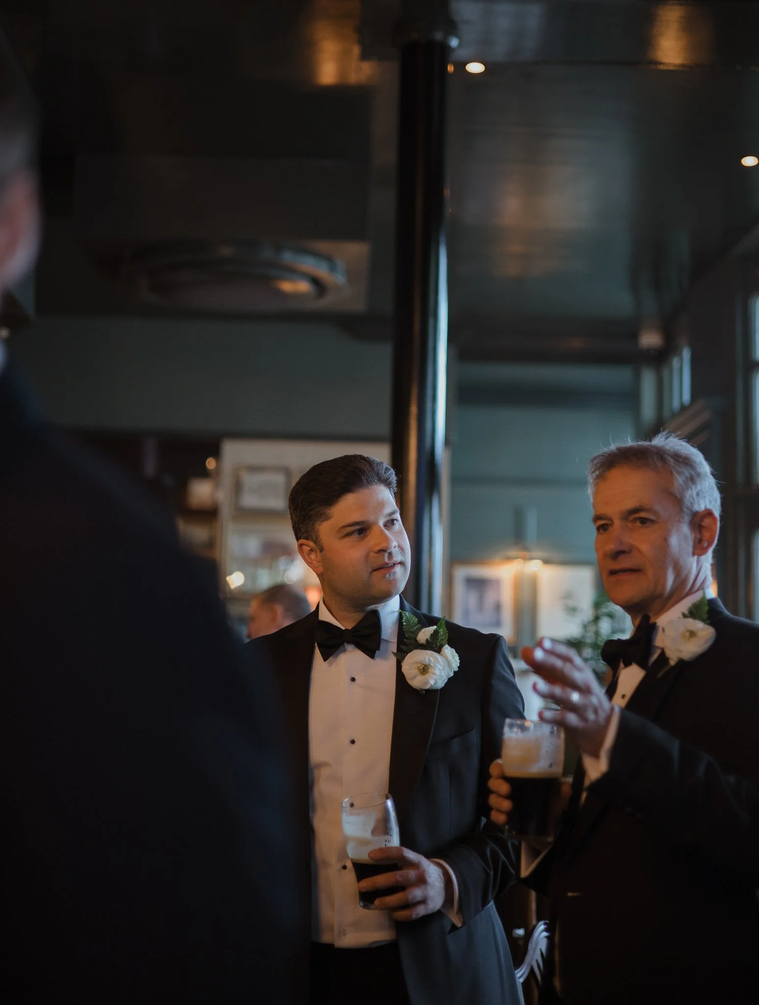 Two men in tuxedos with white floral boutonnieres, holding drinks, engaged in conversation in a dimly lit venue, possibly a wedding reception or formal event.