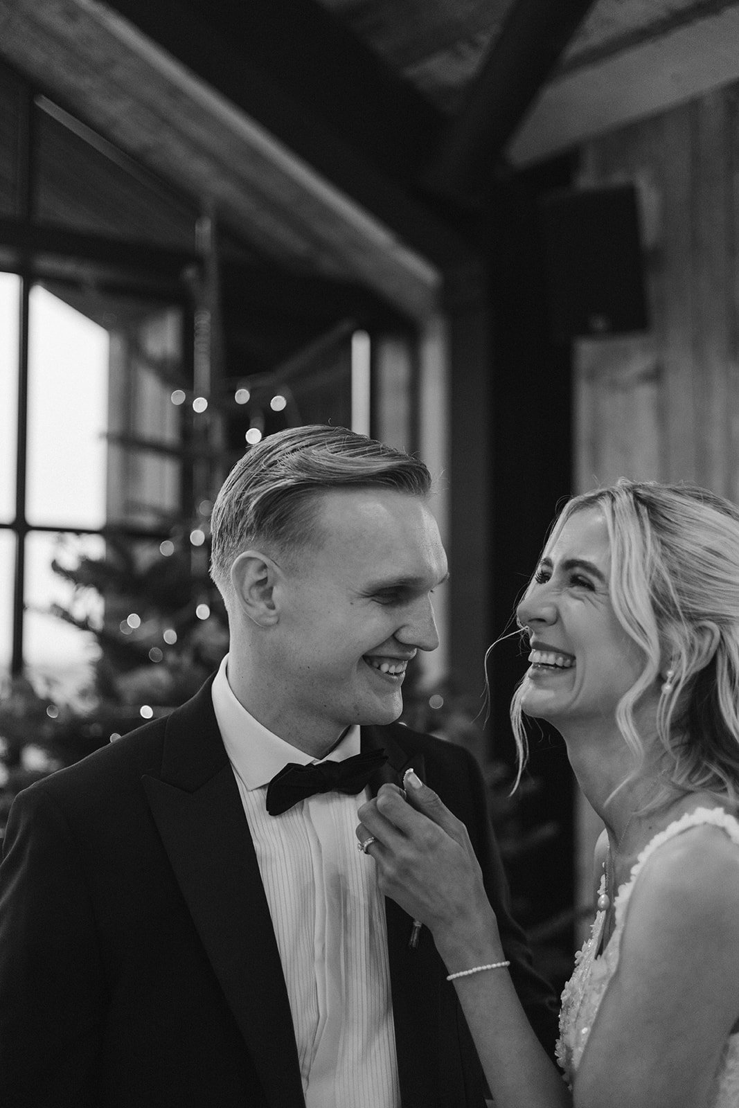 A black and white photo of a smiling bride and groom looking at each other indoors, with Christmas decorations in the background.