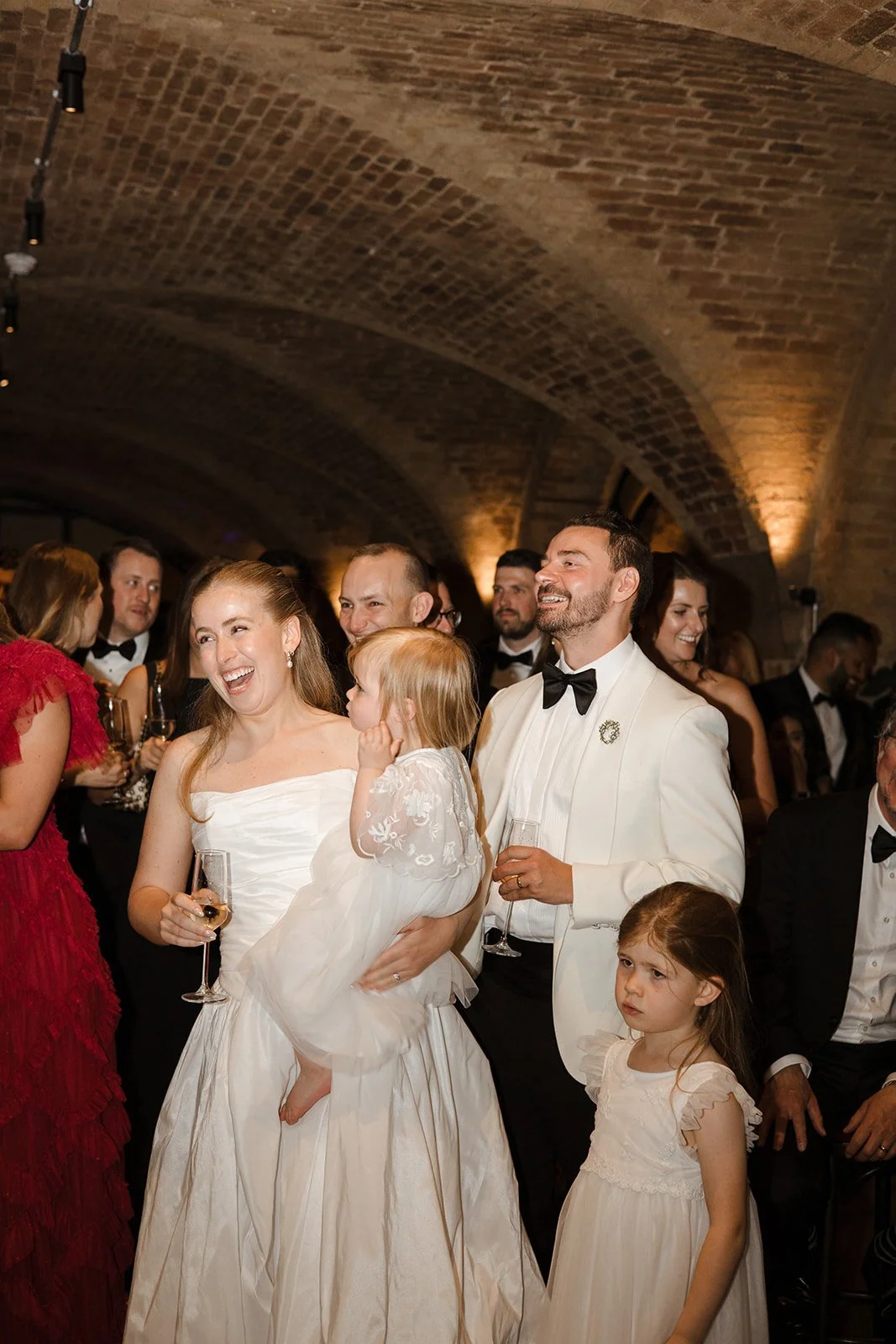 People dressed in formal attire, celebrating at an event with some holding glasses of champagne, inside a brick arched ceiling venue.